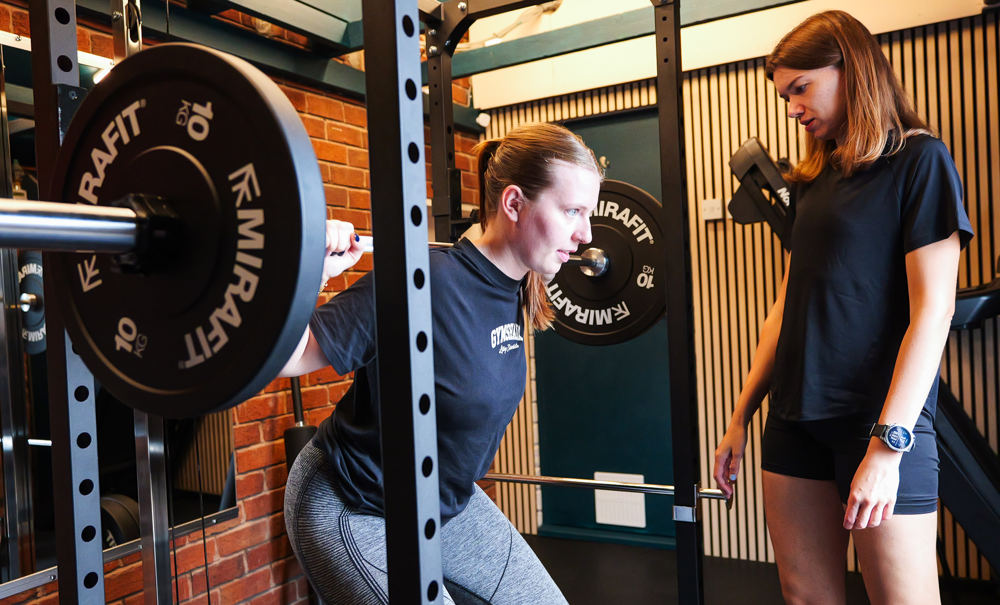 A woman squats with a barbell loaded with 10 kg weights across her shoulders inside a gym, while another woman stands nearby watching.