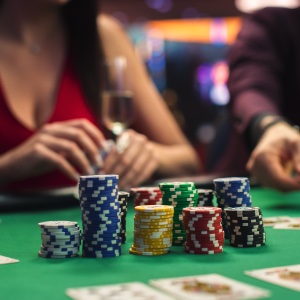An image of a green felt games table with cards and stacks of poker chips on it and people playing in the background