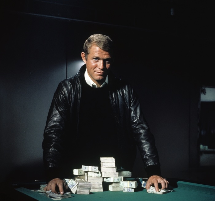 A man standing in a dark room leaning over a green felt topped table with bank notes on it. he is wearing a black jumper, black leather jacket and white collared shirt