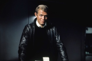 A man standing in a dark room leaning over a green felt topped table with bank notes on it. he is wearing a black jumper, black leather jacket and white collared shirt