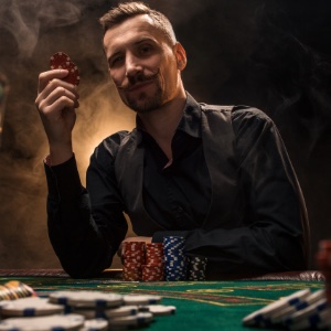 A man wearing a black shirt sitting at a poker table surrounded by cigar smoke and poker chips