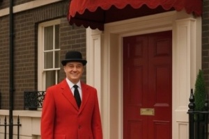 A London terrace house with a man standing outside in a knee length red jacket, a white shirt and black tie with a black bowler hat on outside the front entrance with a red door a canopy above