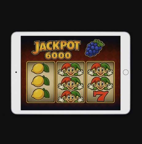 A black background with a silver ipad in the centre, on the screen is an online slot game with fruit and jokers across the reels