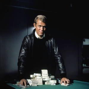 A man standing in a dark room leaning over a green felt topped table with bank notes on it. he is wearing a black jumper, black leather jacket and white collared shirt
