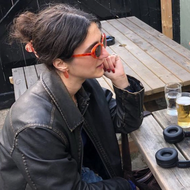 Profile of Marina sat at a table with headphones and a pint on the table. She's wearing red sunglasses. 