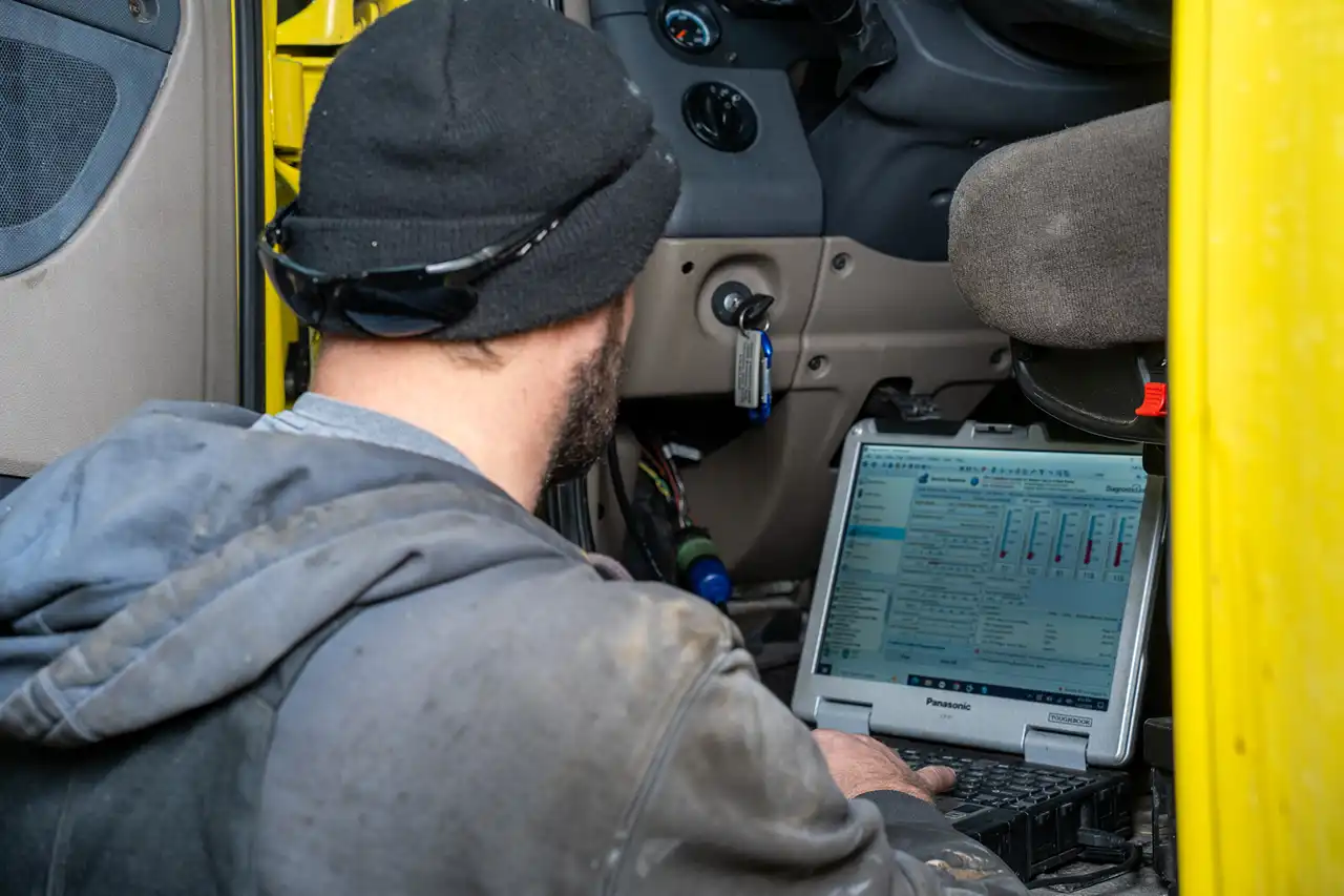 Technician runs diagnostics on a laptop inside a truck cab, checking engine performance data and fault codes.