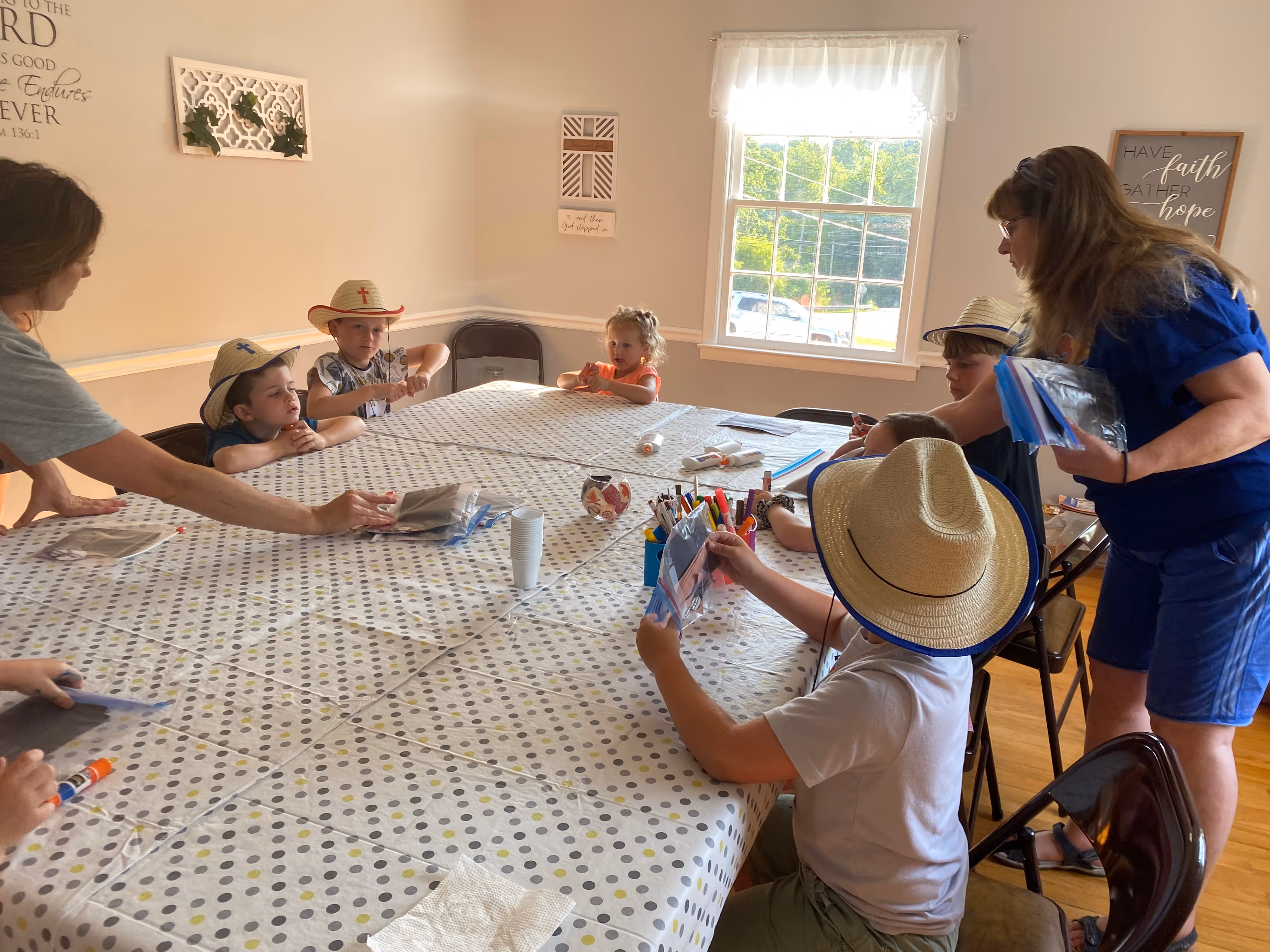 Children wearing straw hats with crosses sit around a large table covered with a polka dot tablecloth, while two adults distribute craft supplies in a sunlit room.