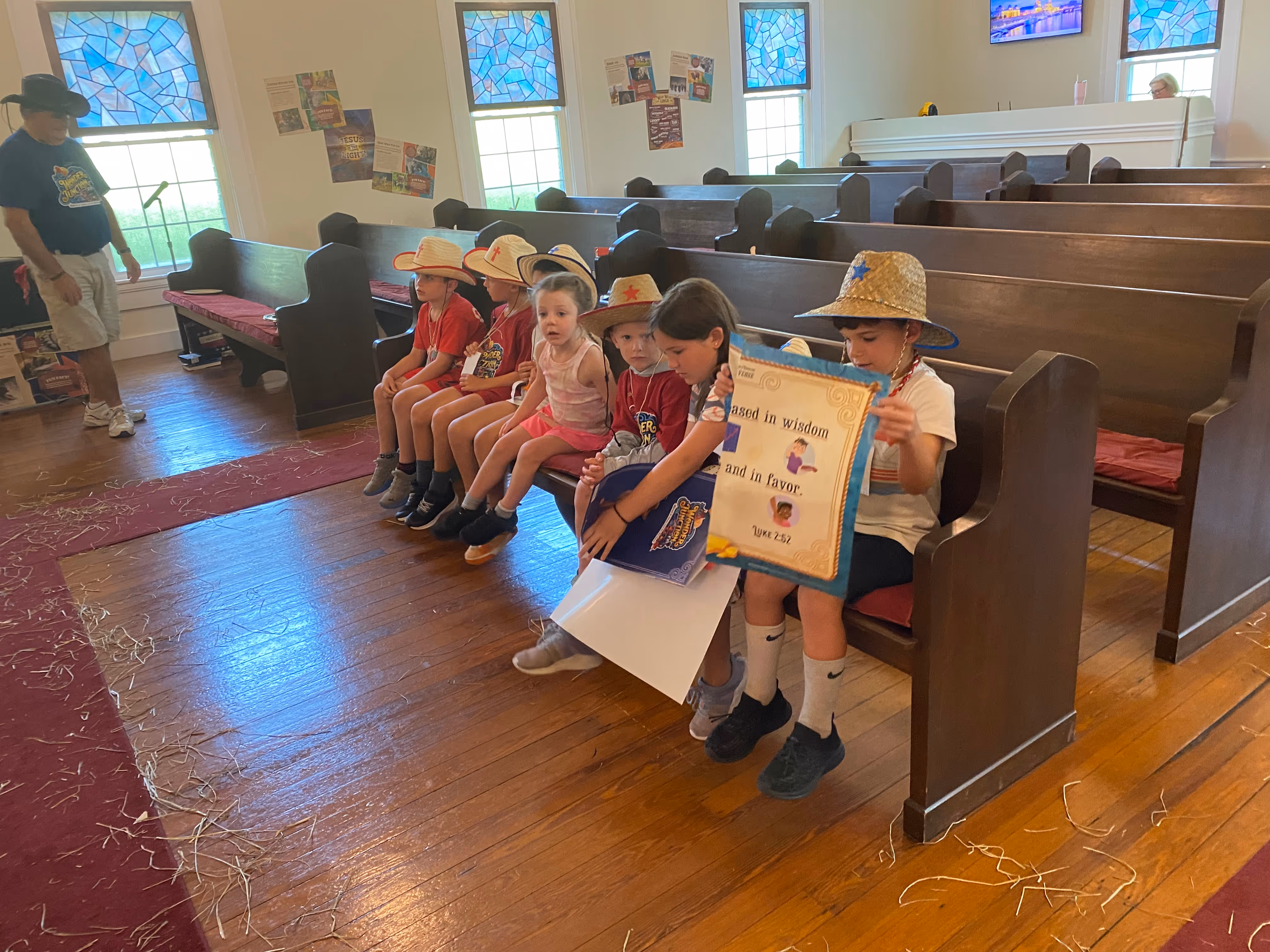 Group of children wearing hats sitting on a church pew, one holding a sign with a biblical quote.