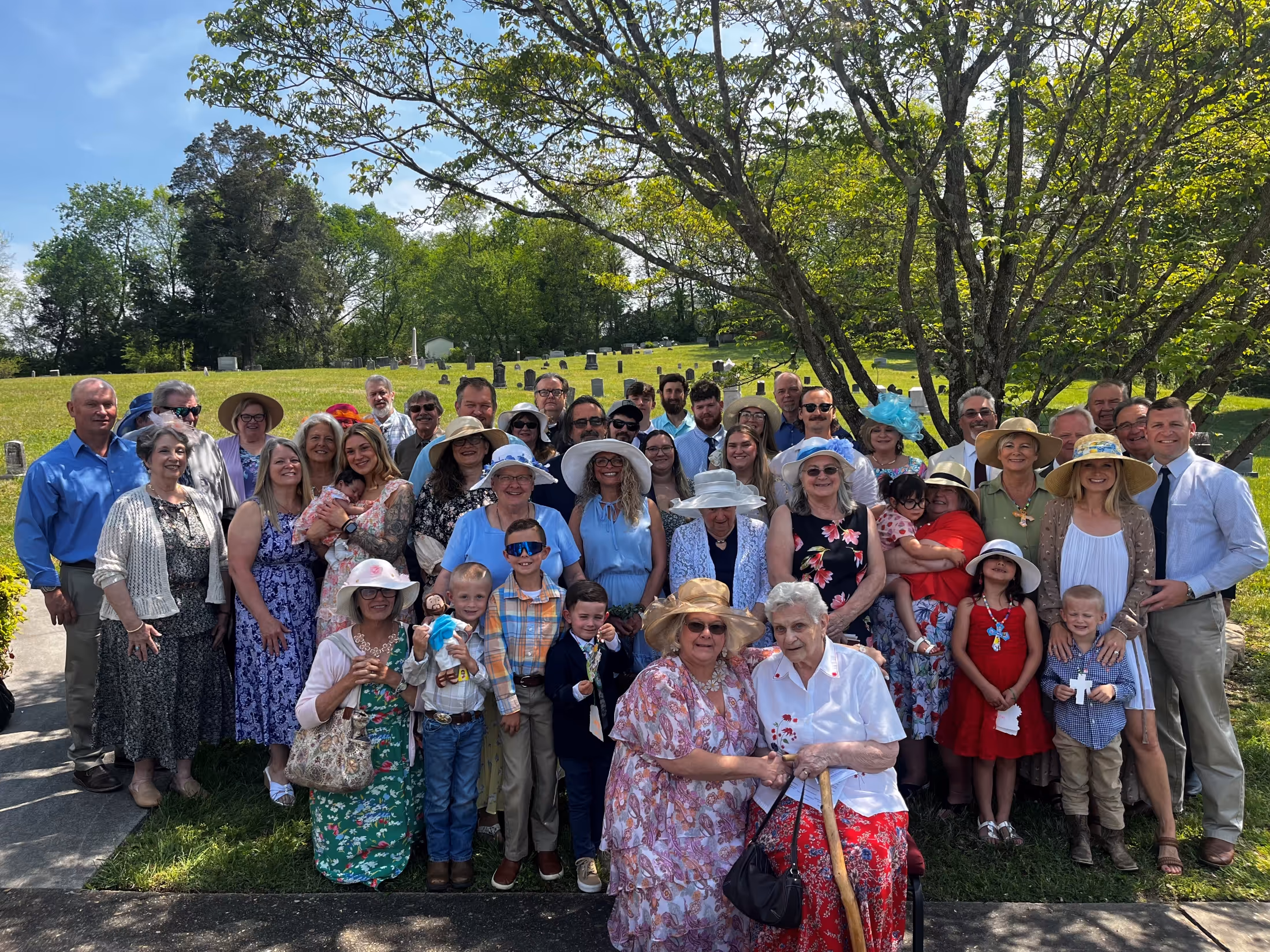Large family group of multiple generations gathered outdoors on a sunny day, with some women wearing hats and a cemetery visible in the background.