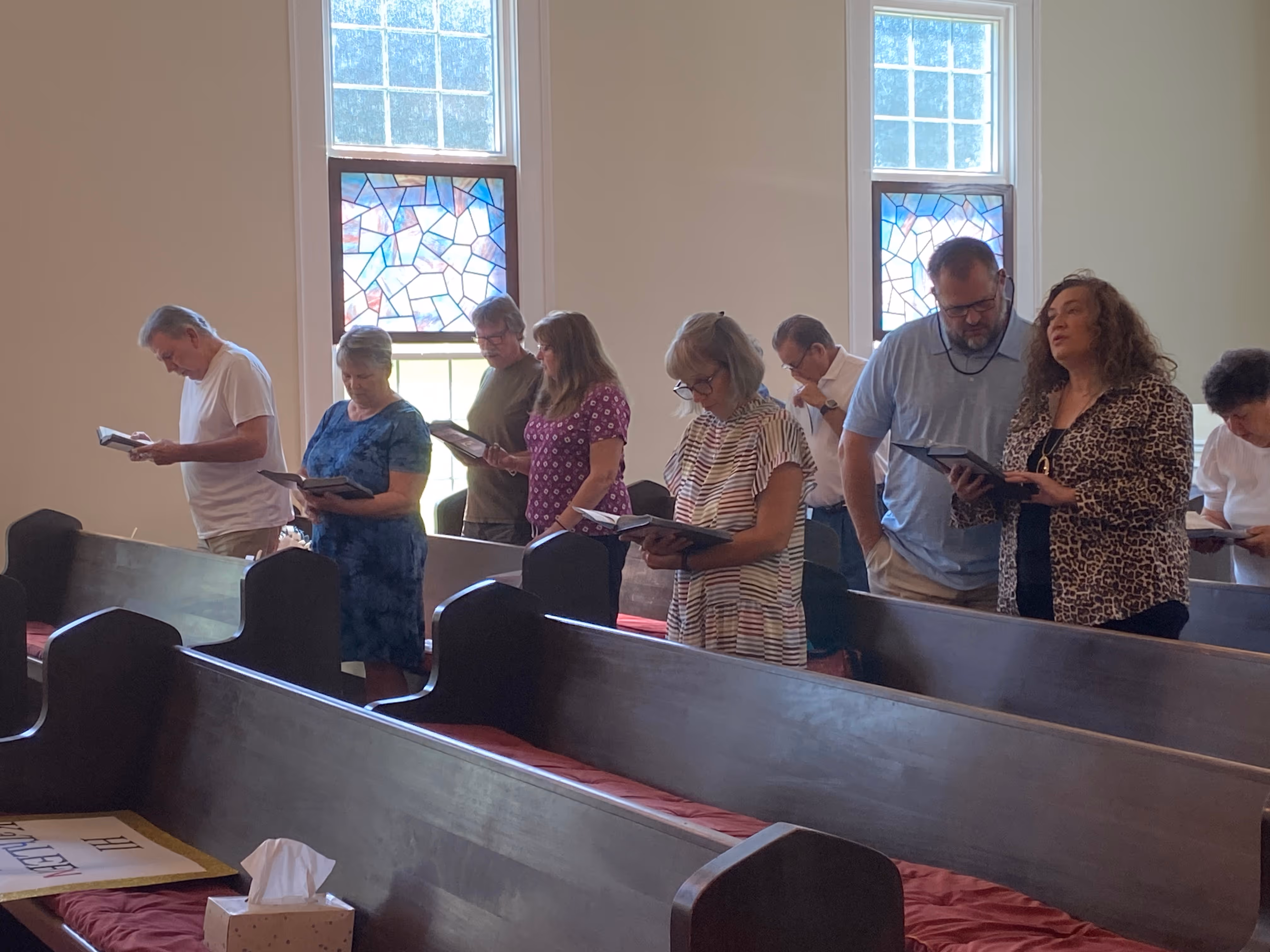Group of people standing in church pews reading hymnals with stained glass windows in the background.