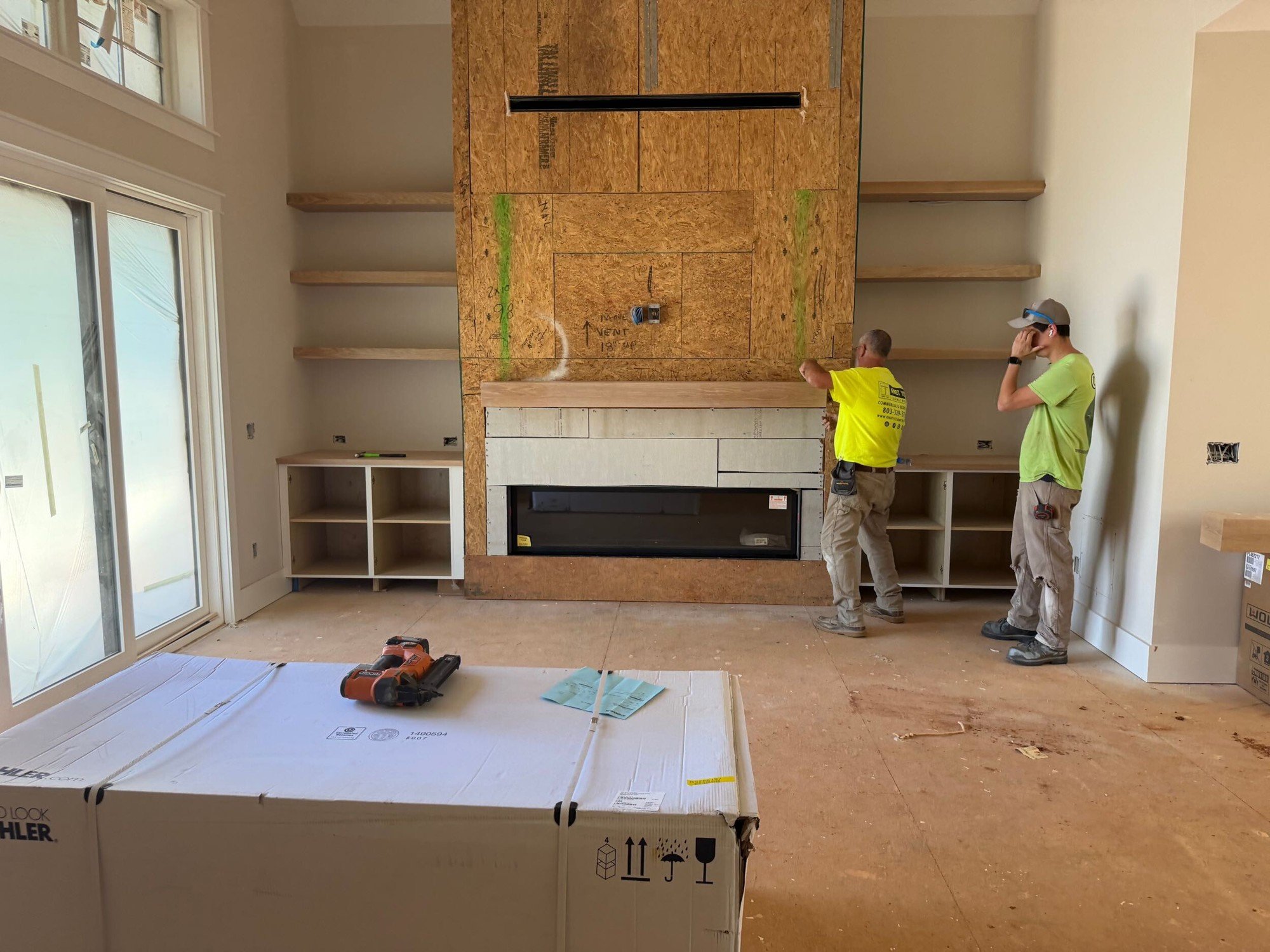 Fireplace wall before stone, with plywood backer, a black linear firebox, a wood mantel and built-in cabinets on each side.