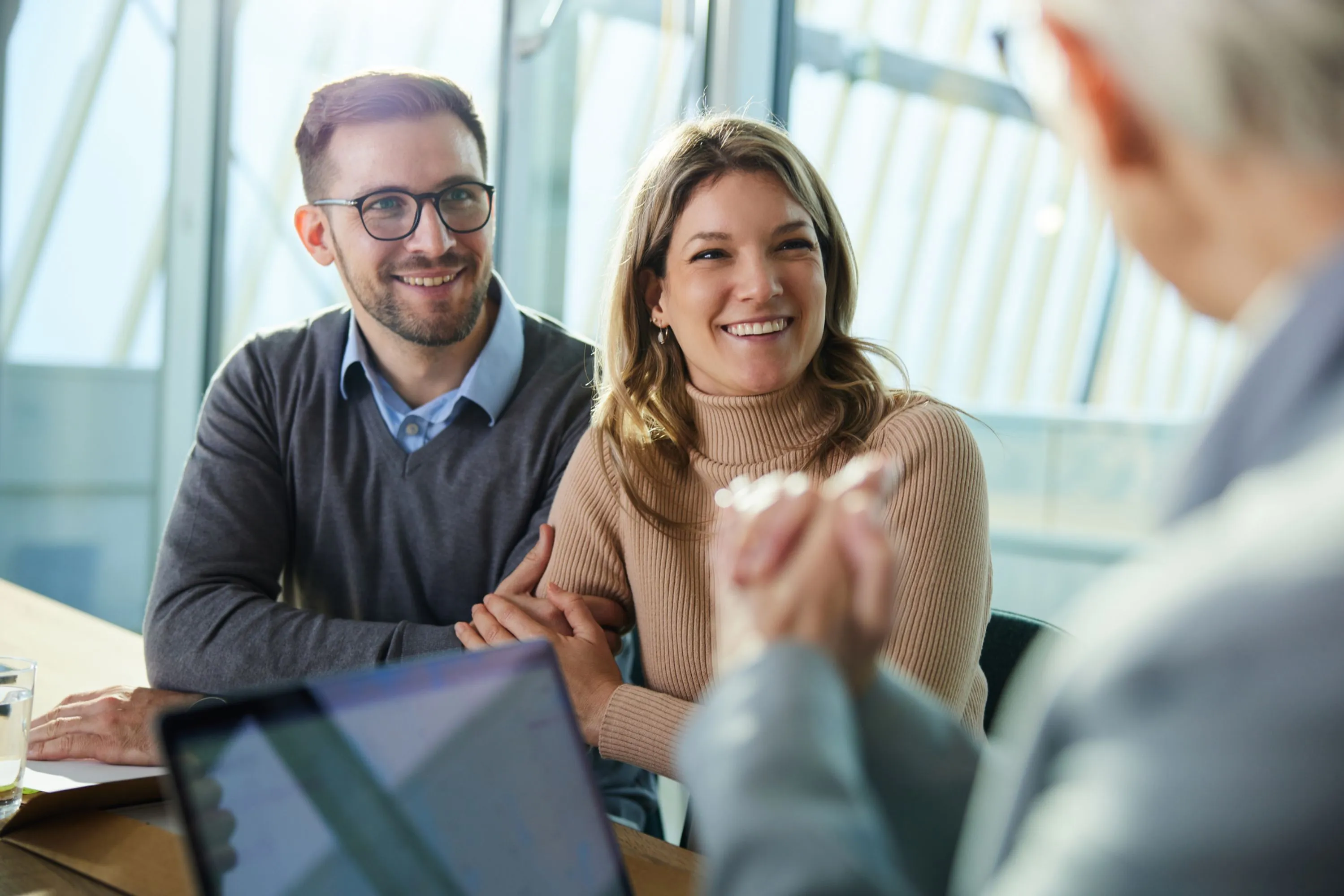 Smiling young couple sitting together at a table, attentively listening to a person in the foreground.