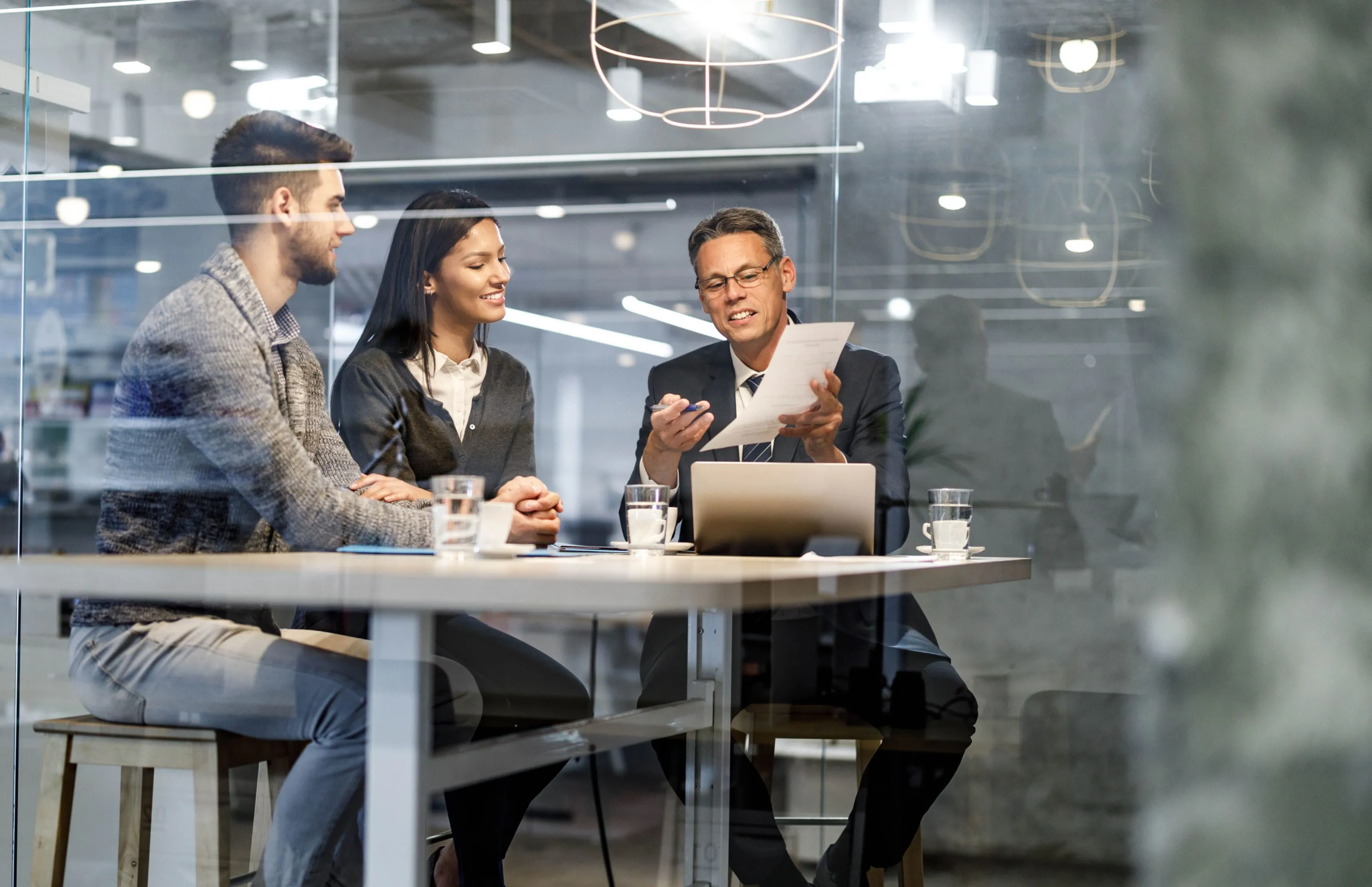 Three business professionals sitting at a table in a modern office, reviewing documents together.