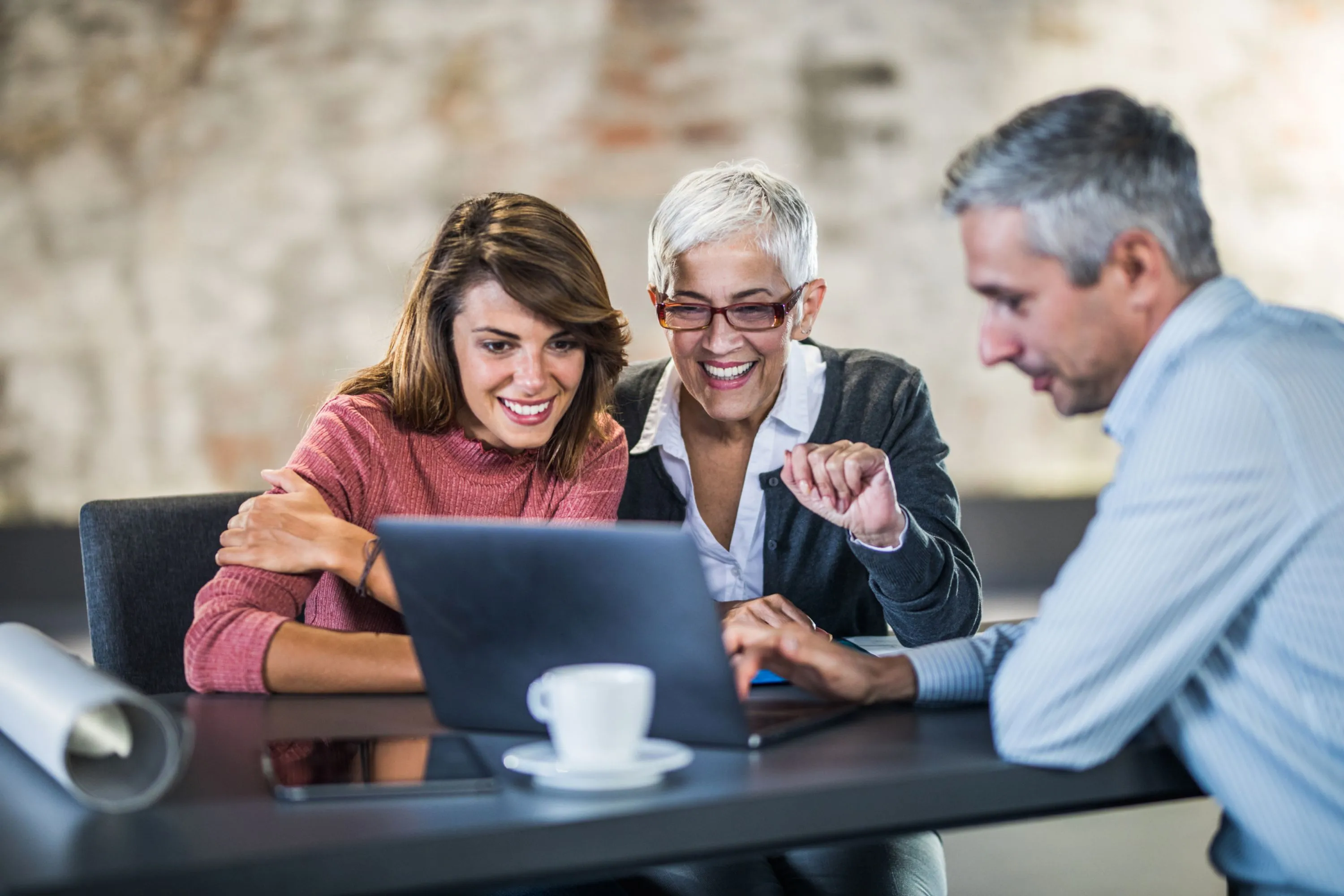 Three colleagues smiling and looking at a laptop screen together in a casual office setting.