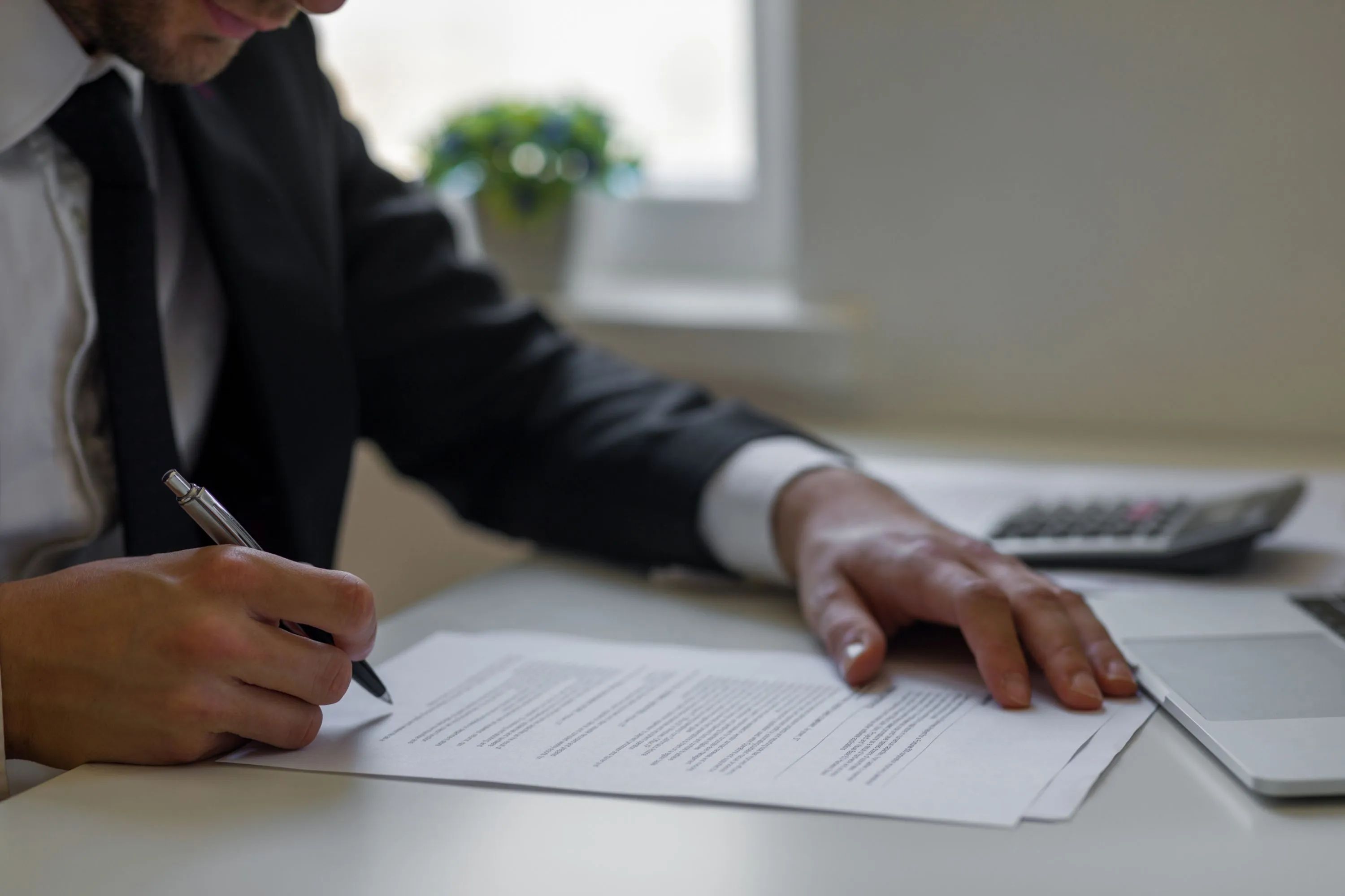 Person in a black suit signing a document on a desk near a laptop and calculator.