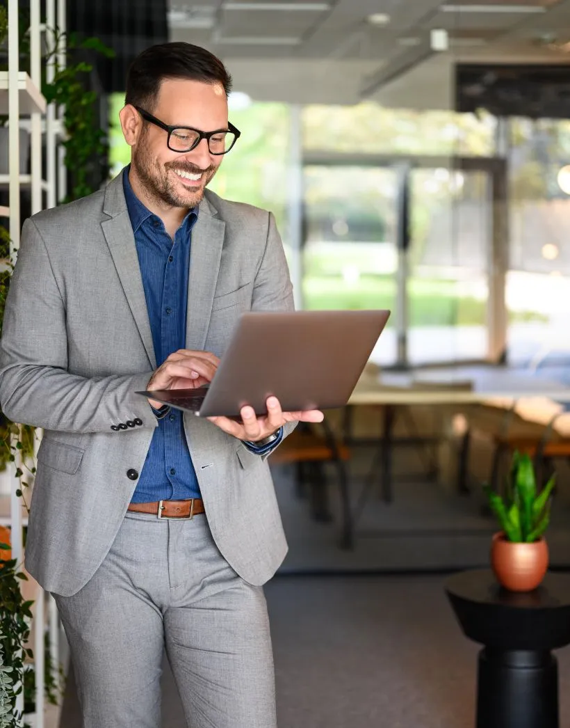 Smiling man in gray suit and blue shirt using a laptop in a bright office with plants.