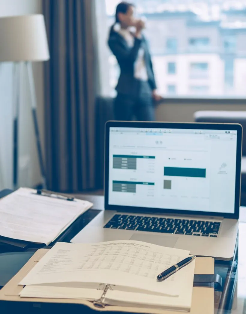 Desk with open binder, pen, clipboard, and laptop showing charts, with a businesswoman drinking coffee by a window in the background.