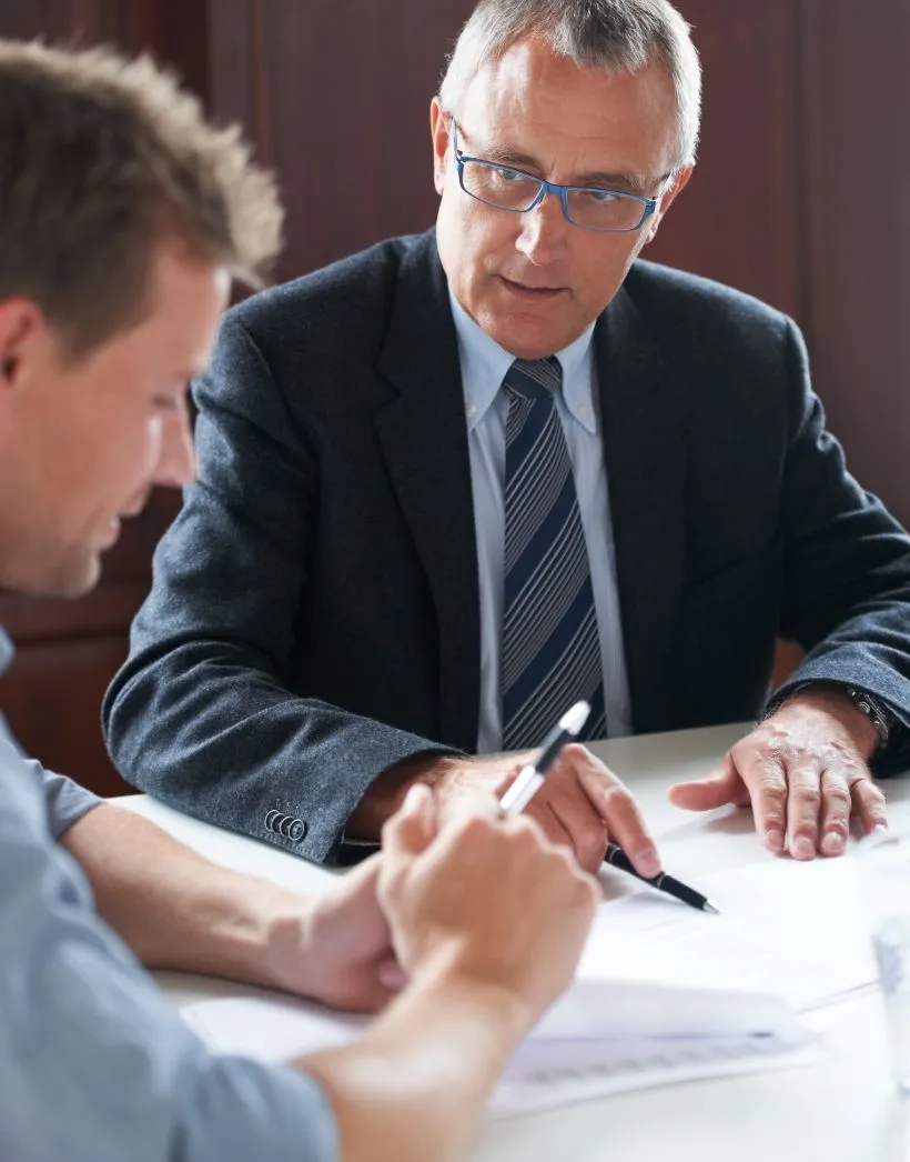 Two men in a business meeting, one reviewing documents and the other pointing at papers on a table.