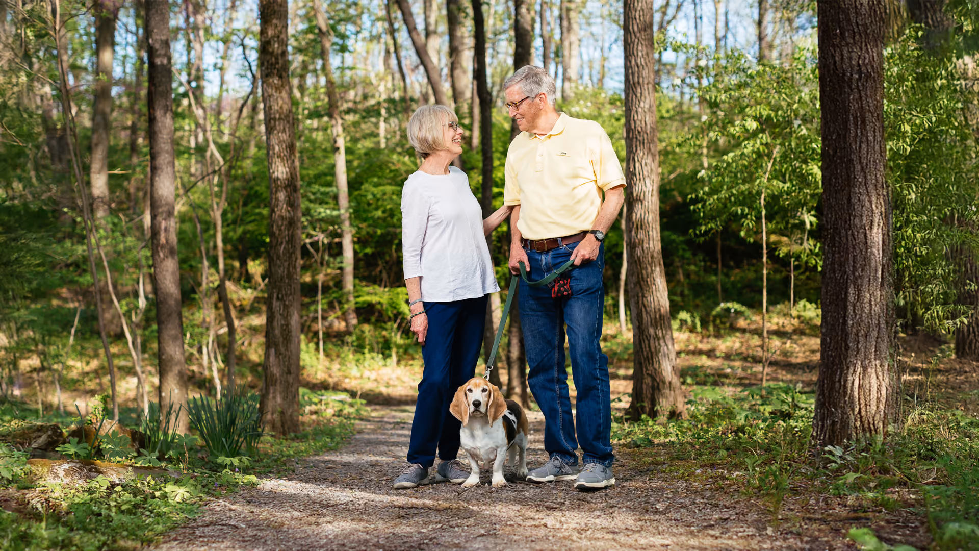 An elderly couple walks with their beagle dog on a forest path, smiling at each other.