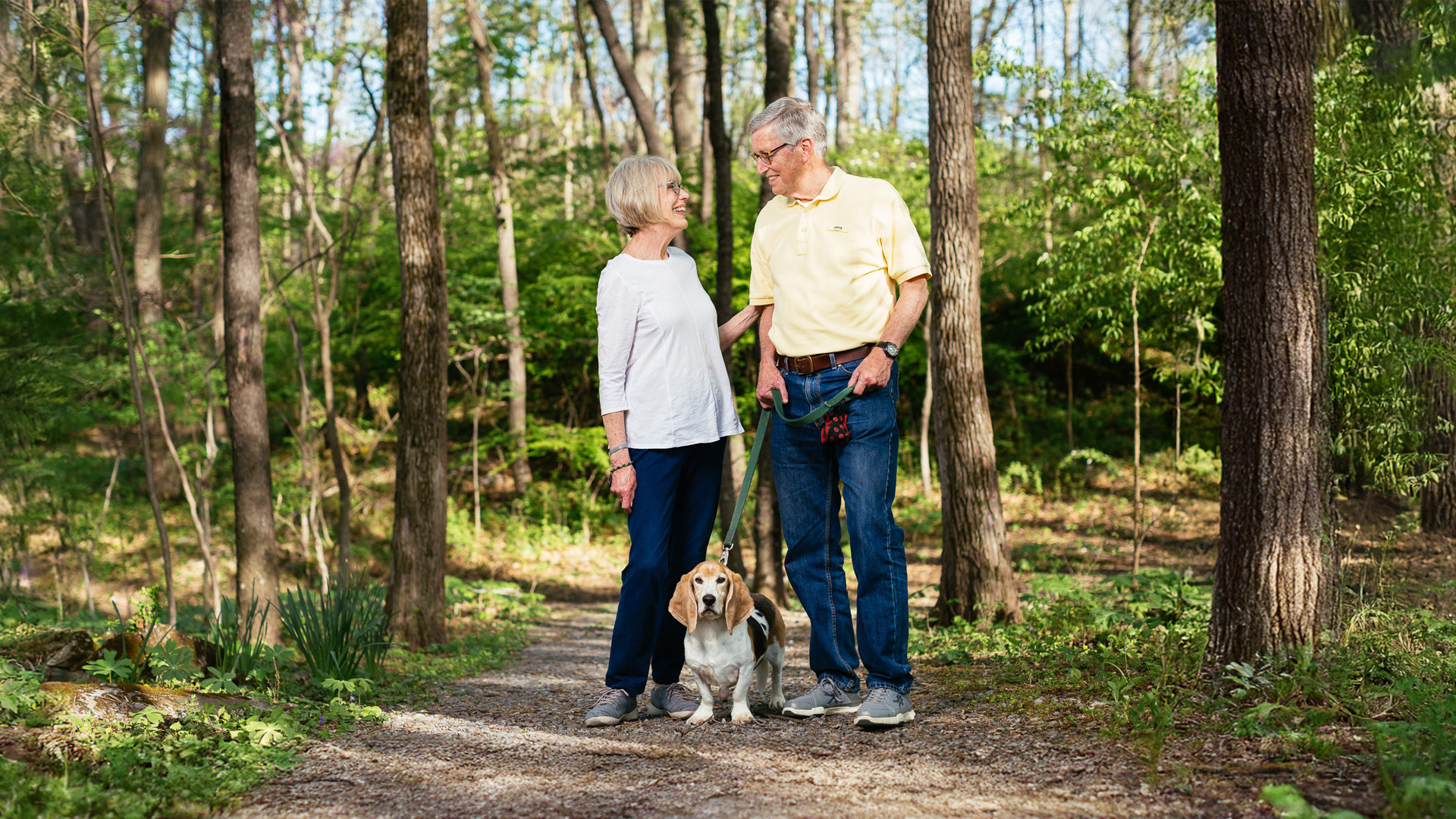 An elderly couple walks with their beagle dog on a forest path, smiling at each other.