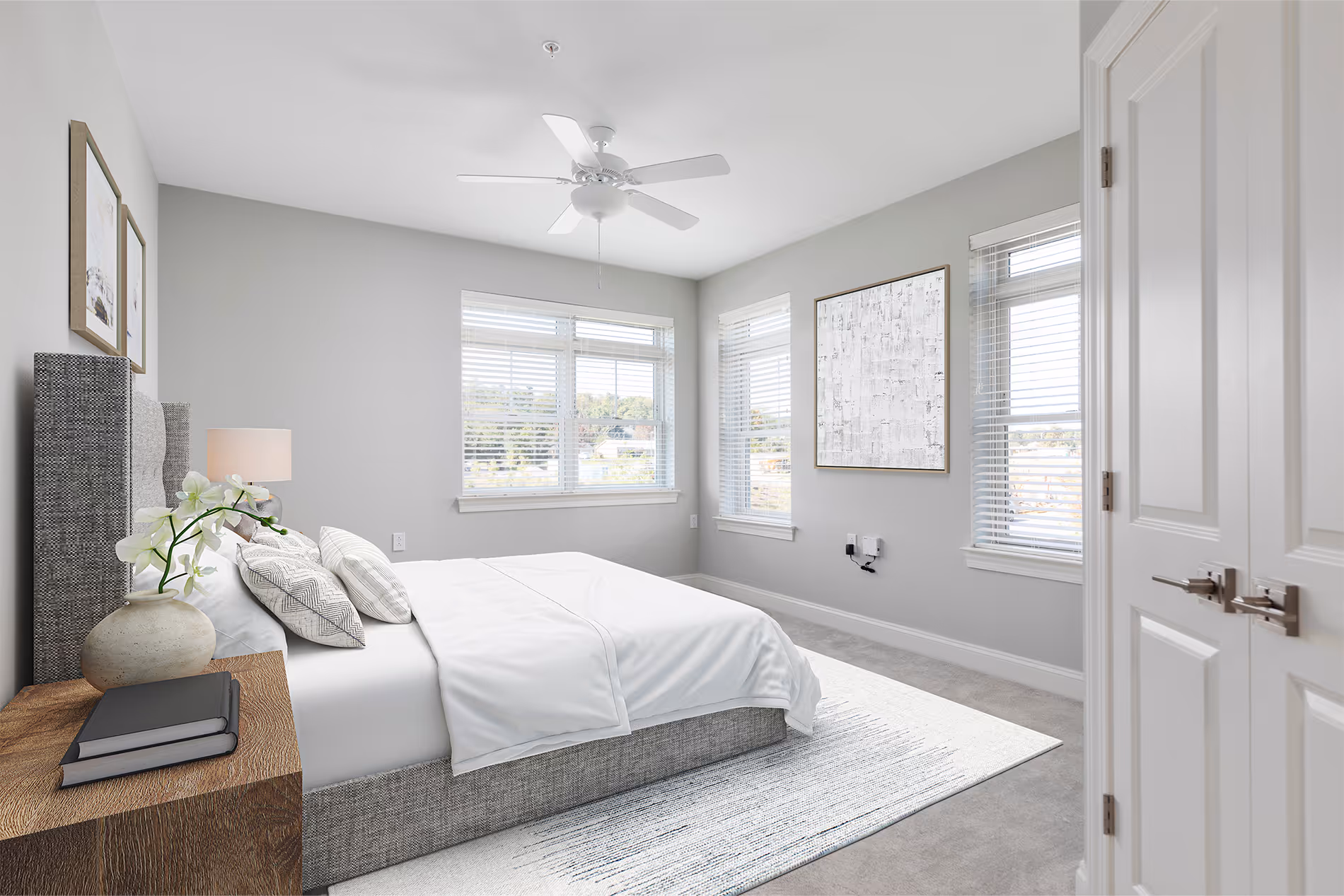A neatly made bed in a bright bedroom with gray walls and large windows showing natural light.