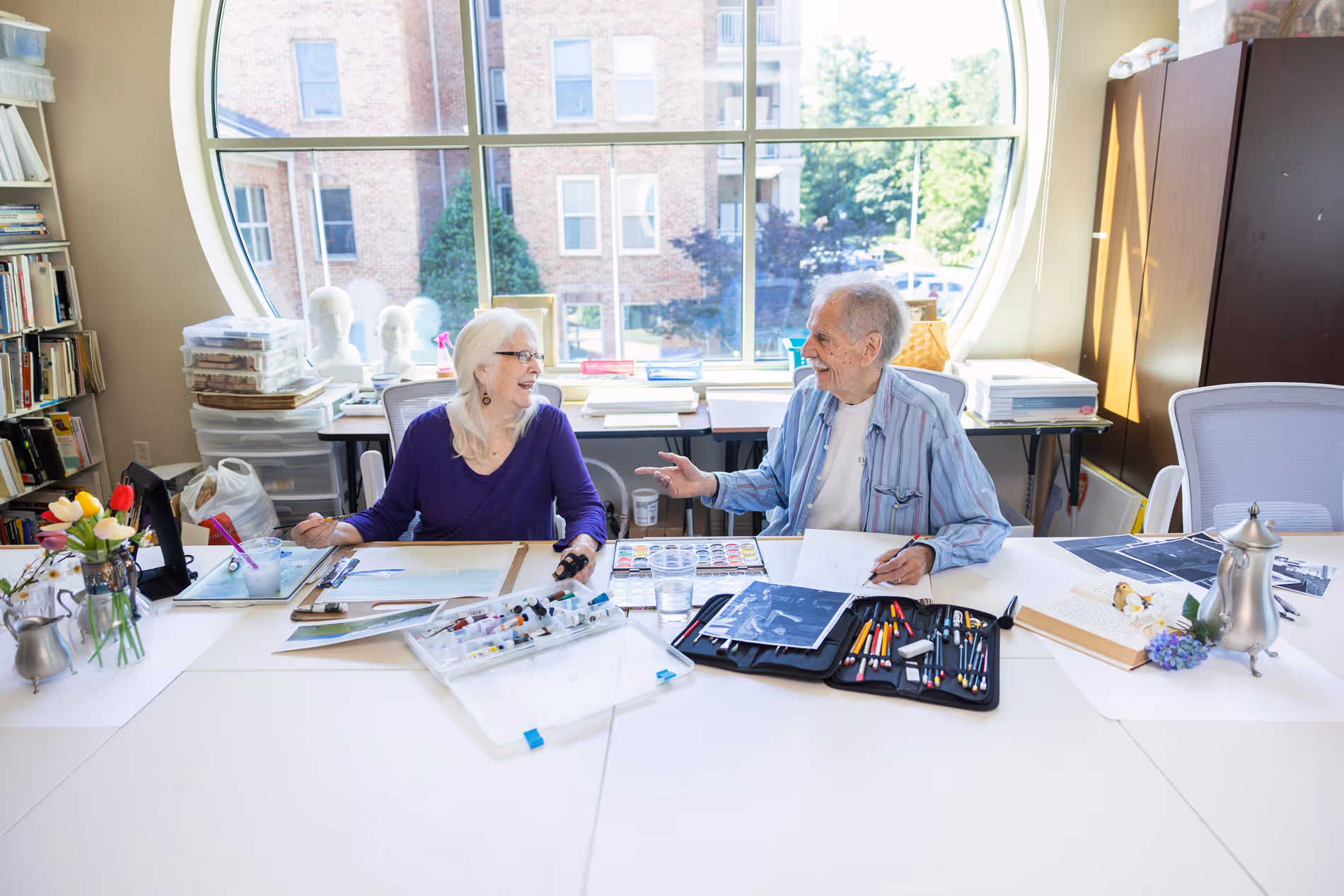 Two people sit at a table with art supplies, smiling and talking to each other.