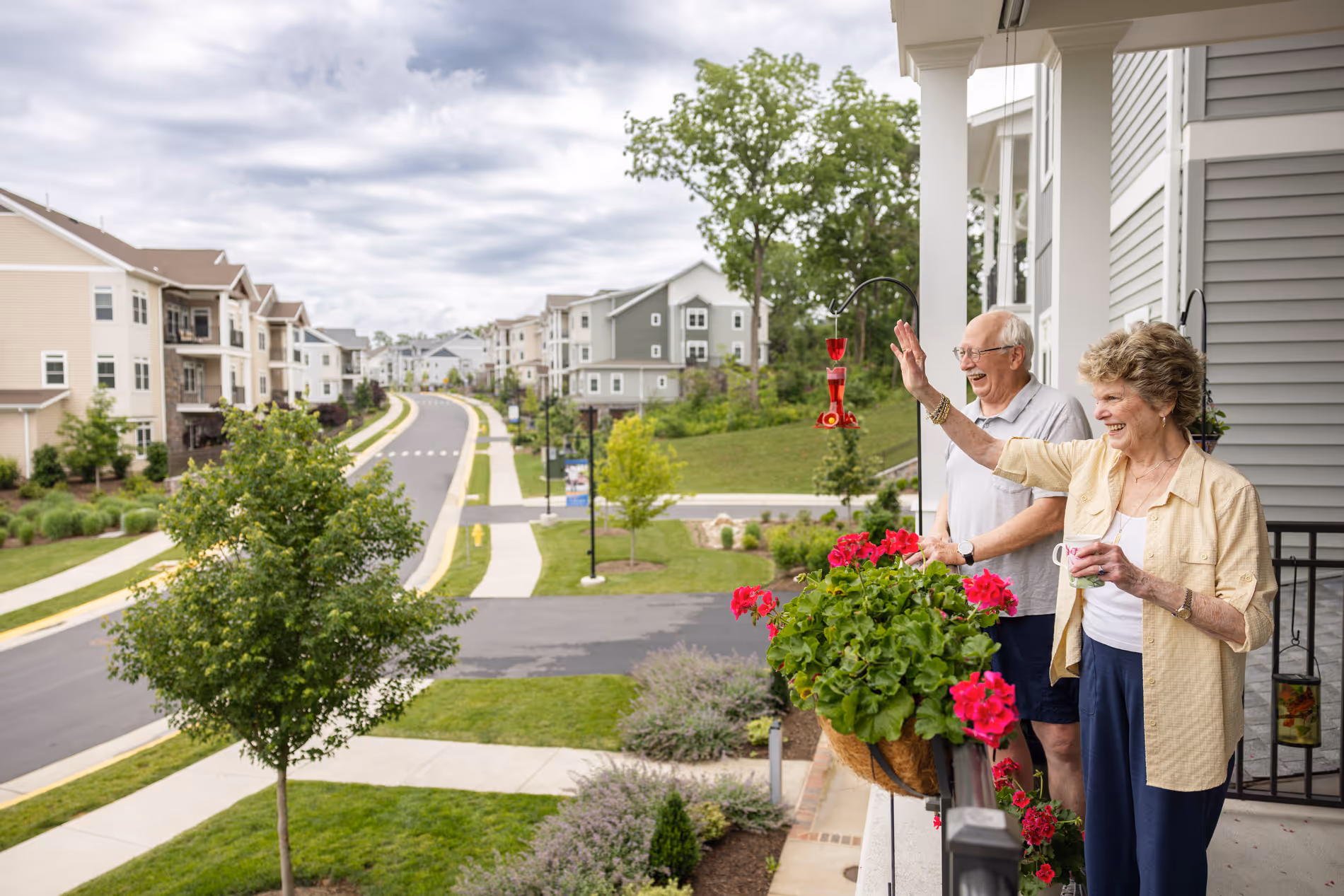 An elderly couple stands on a balcony, waving to someone, surrounded by suburban houses and greenery.