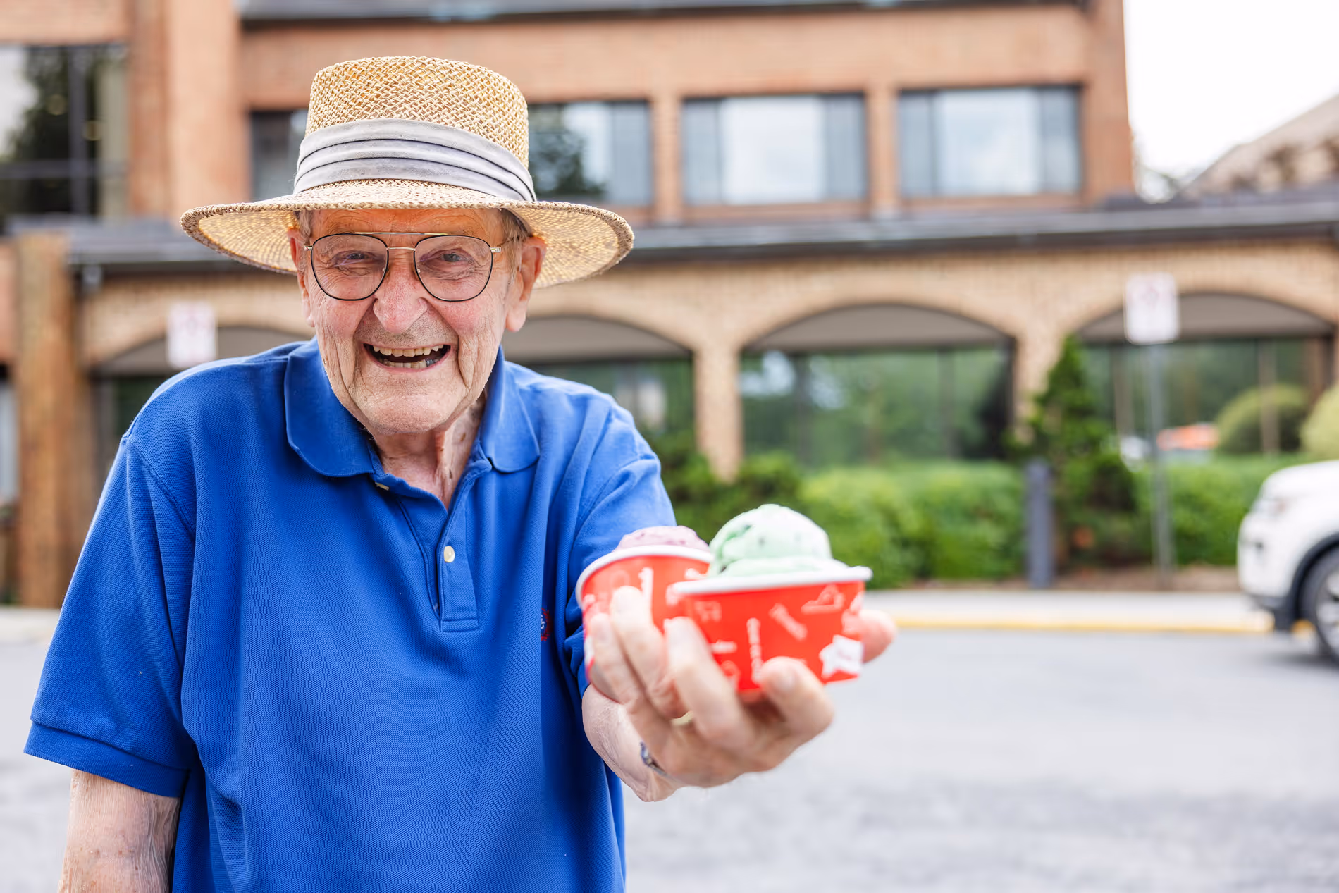 Elderly man wearing a straw hat offers two cups of ice cream with a smile.