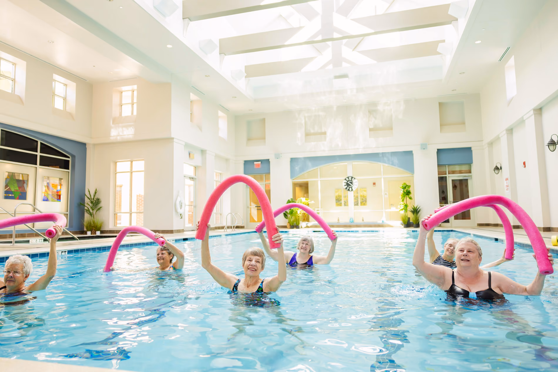 A group of elderly people exercise with pool noodles in an indoor swimming pool.