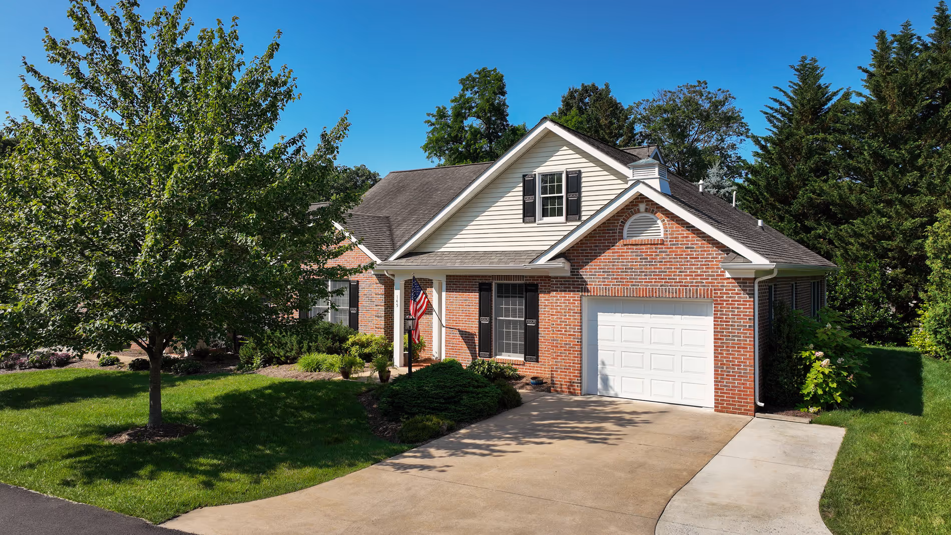 Single-story brick house with white garage door, front porch displaying an American flag, and a large tree on the lawn.