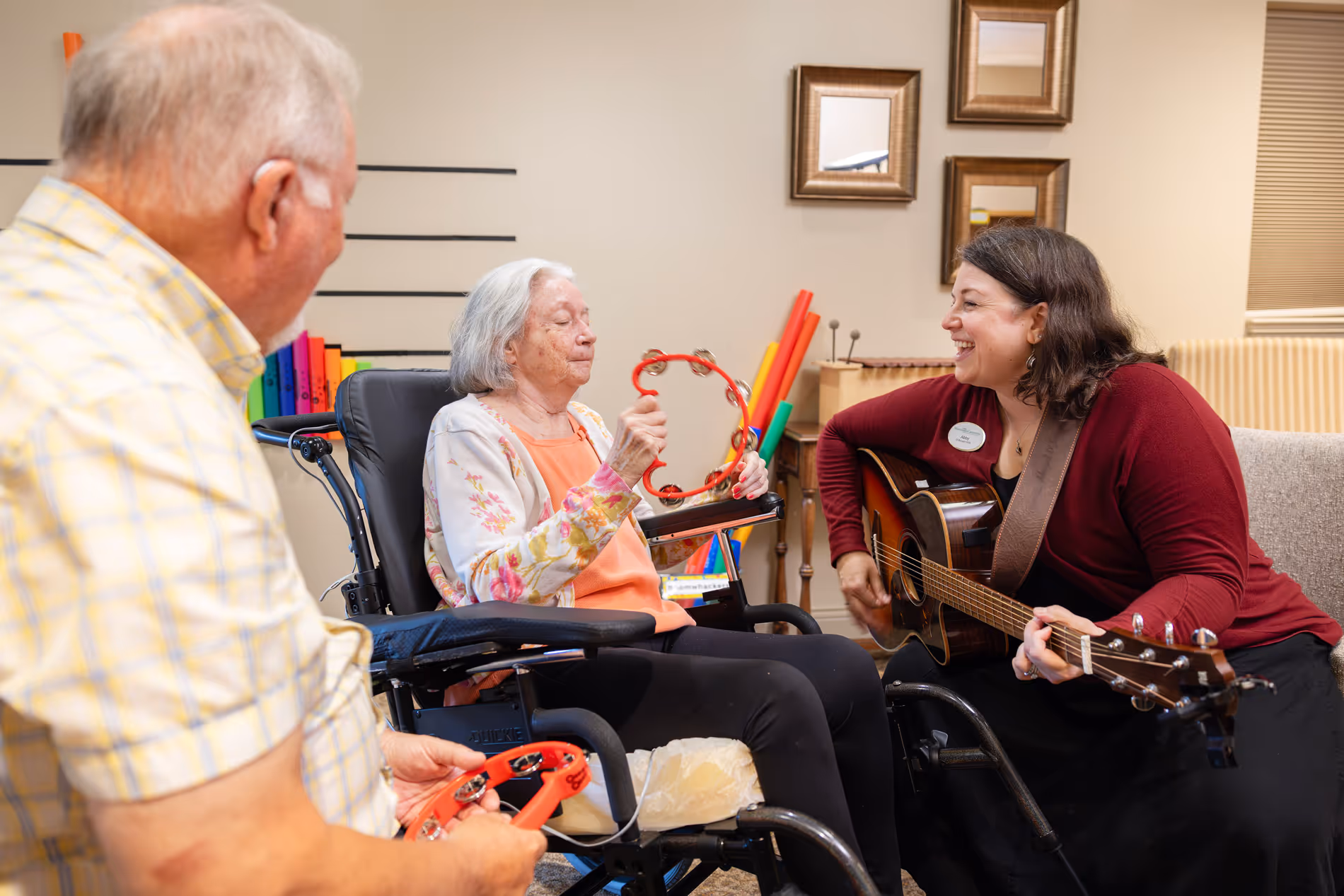An elderly woman in a wheelchair plays a tambourine alongside a woman playing a guitar.