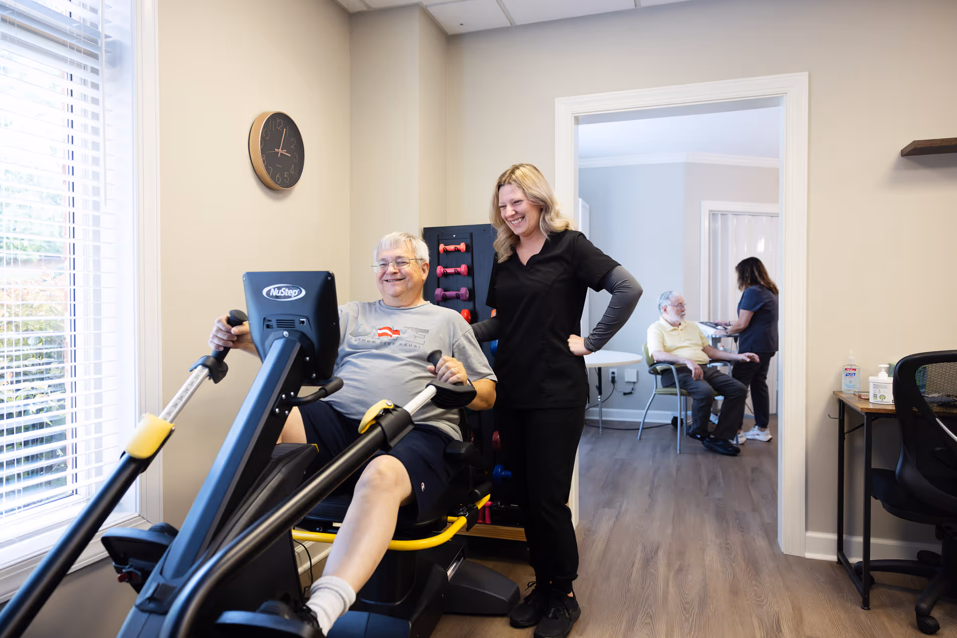 A man exercises on a stationary bike while a woman stands beside him, smiling.