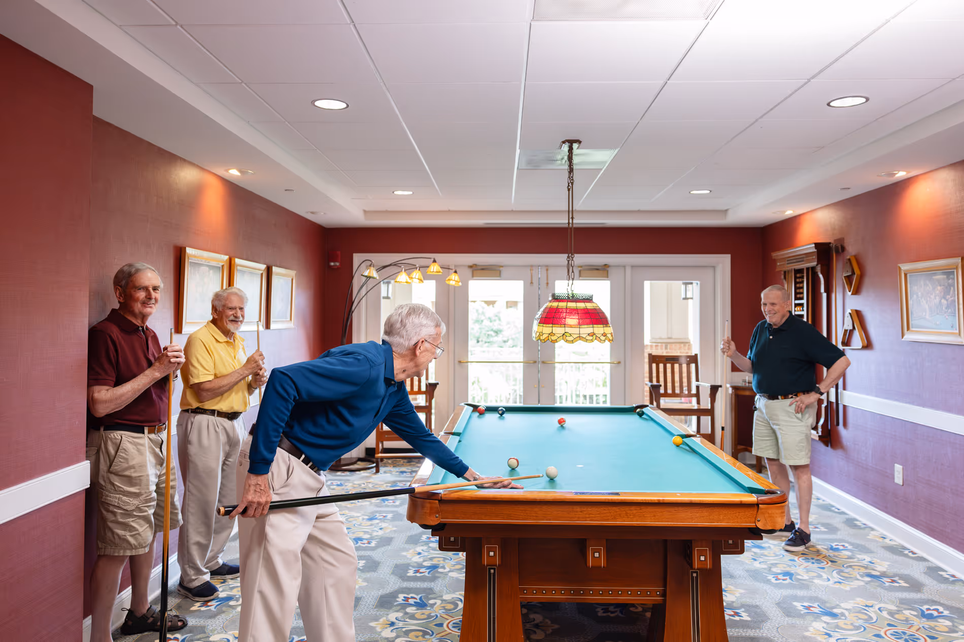 Four men are playing pool in a room with a red wall and a large window.