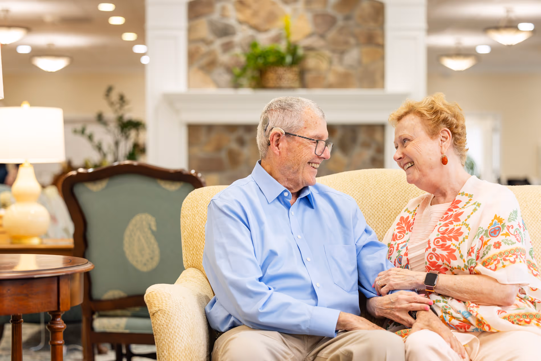 Elderly couple sitting on a sofa, smiling and holding hands in a warmly lit room.