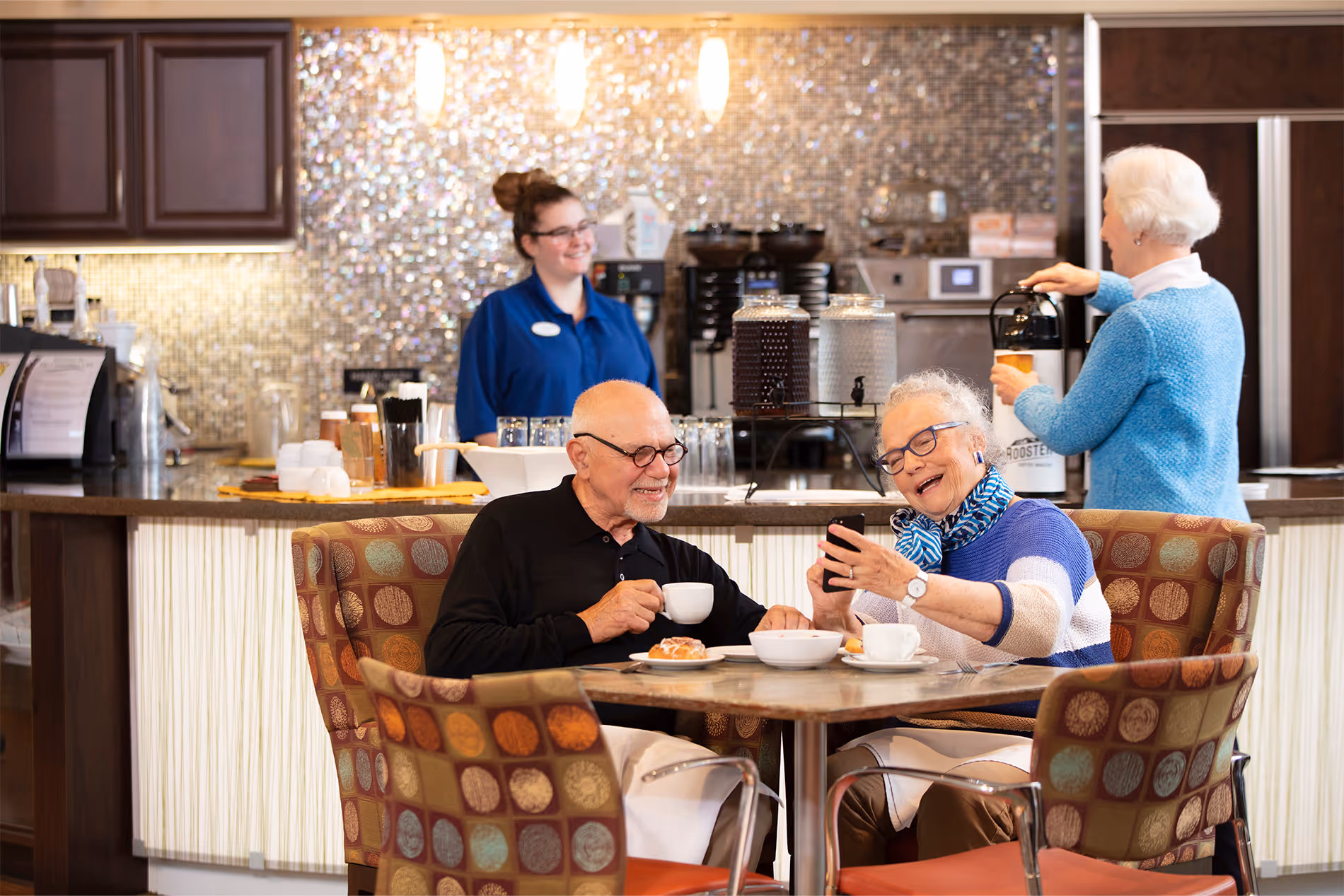 Two older adults at a café table smile while looking at a smartphone, with coffee and pastries.