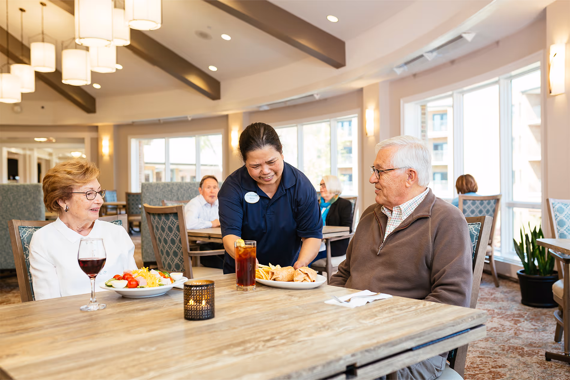 A server delivers plates of food to an older couple seated at a dining table.