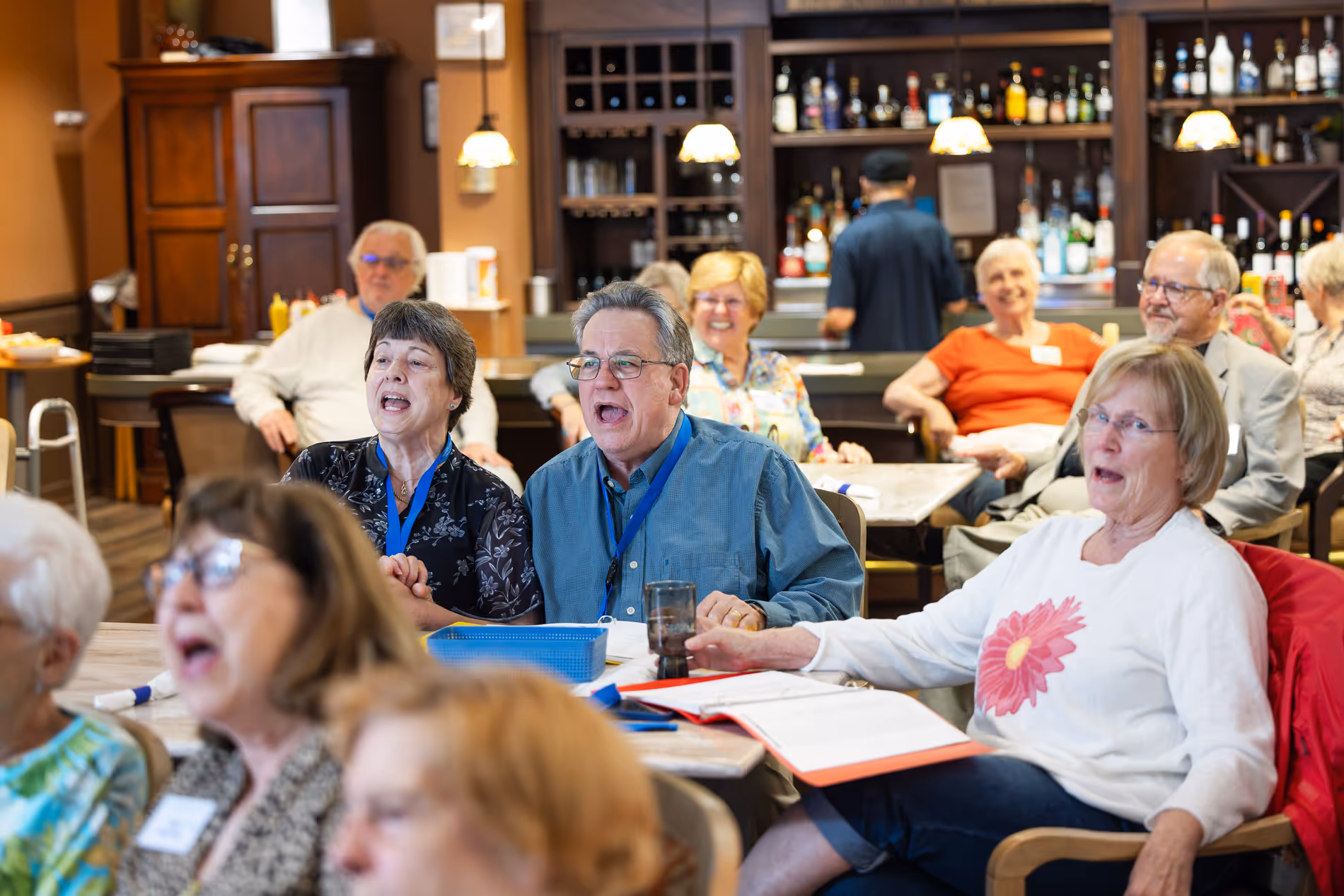 A group of older adults sits together enthusiastically singing at a social gathering indoors.