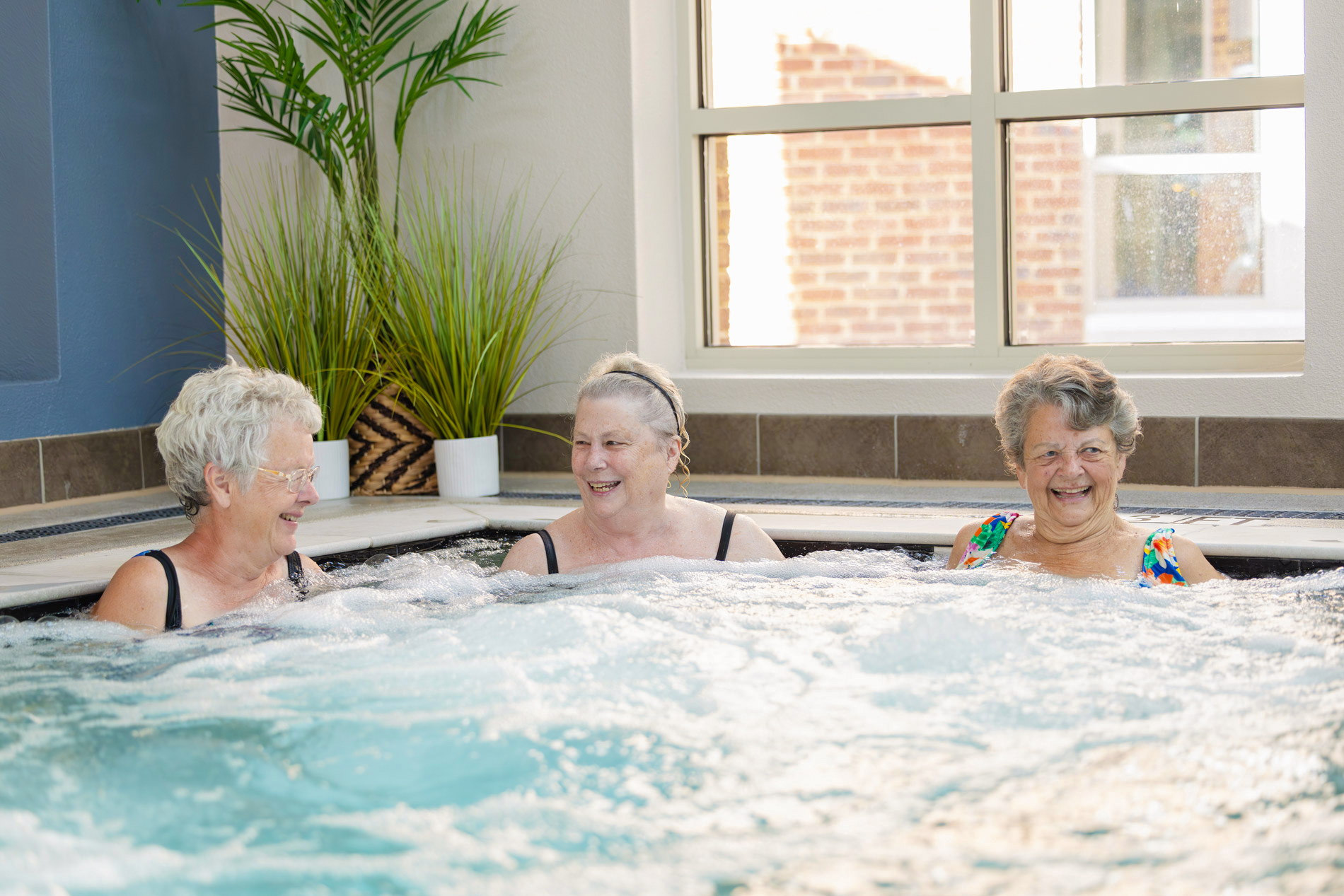 Three elderly women sitting together and smiling while relaxing in a hot tub indoors.