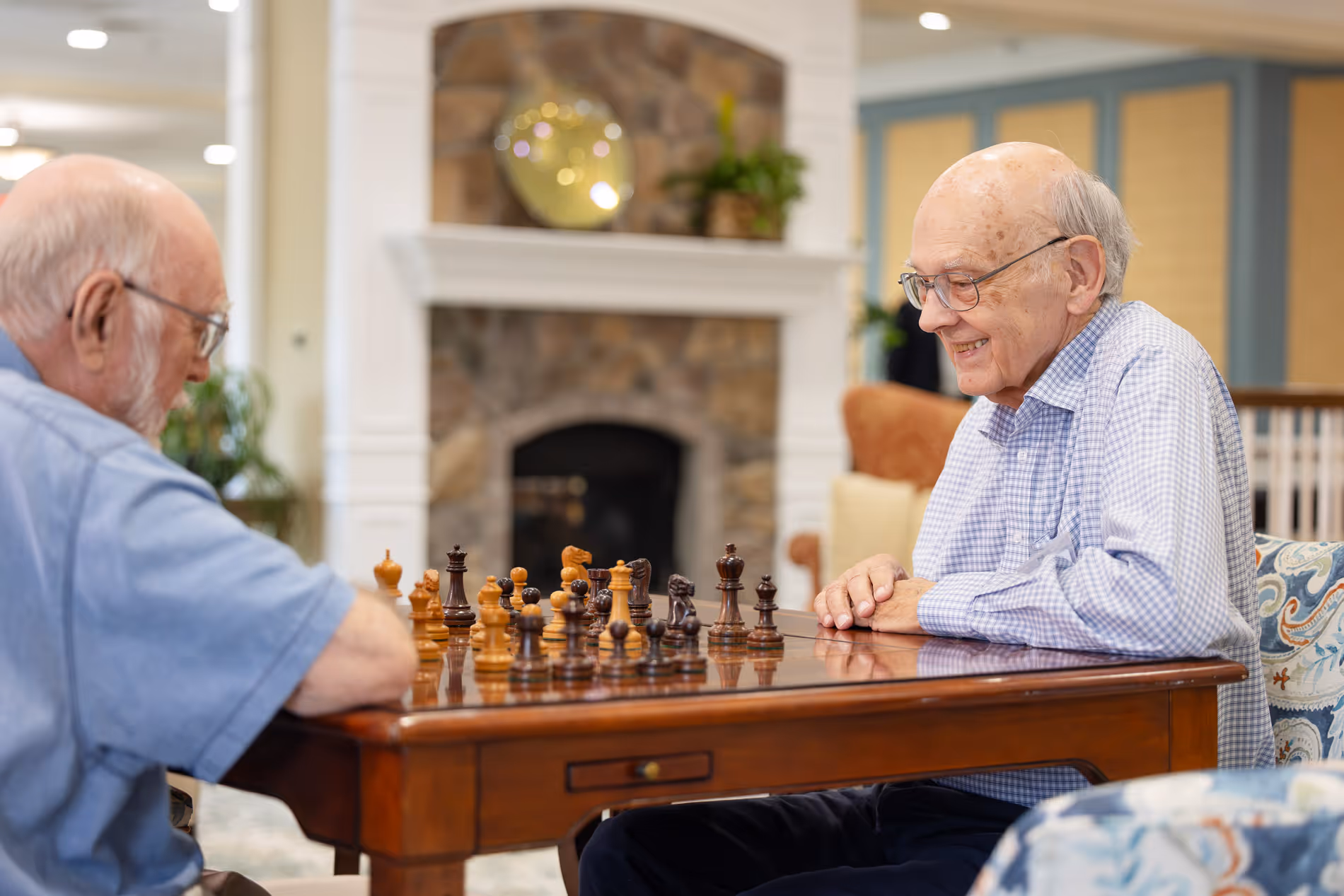 Two elderly men play chess at a wooden table in a cozy room.