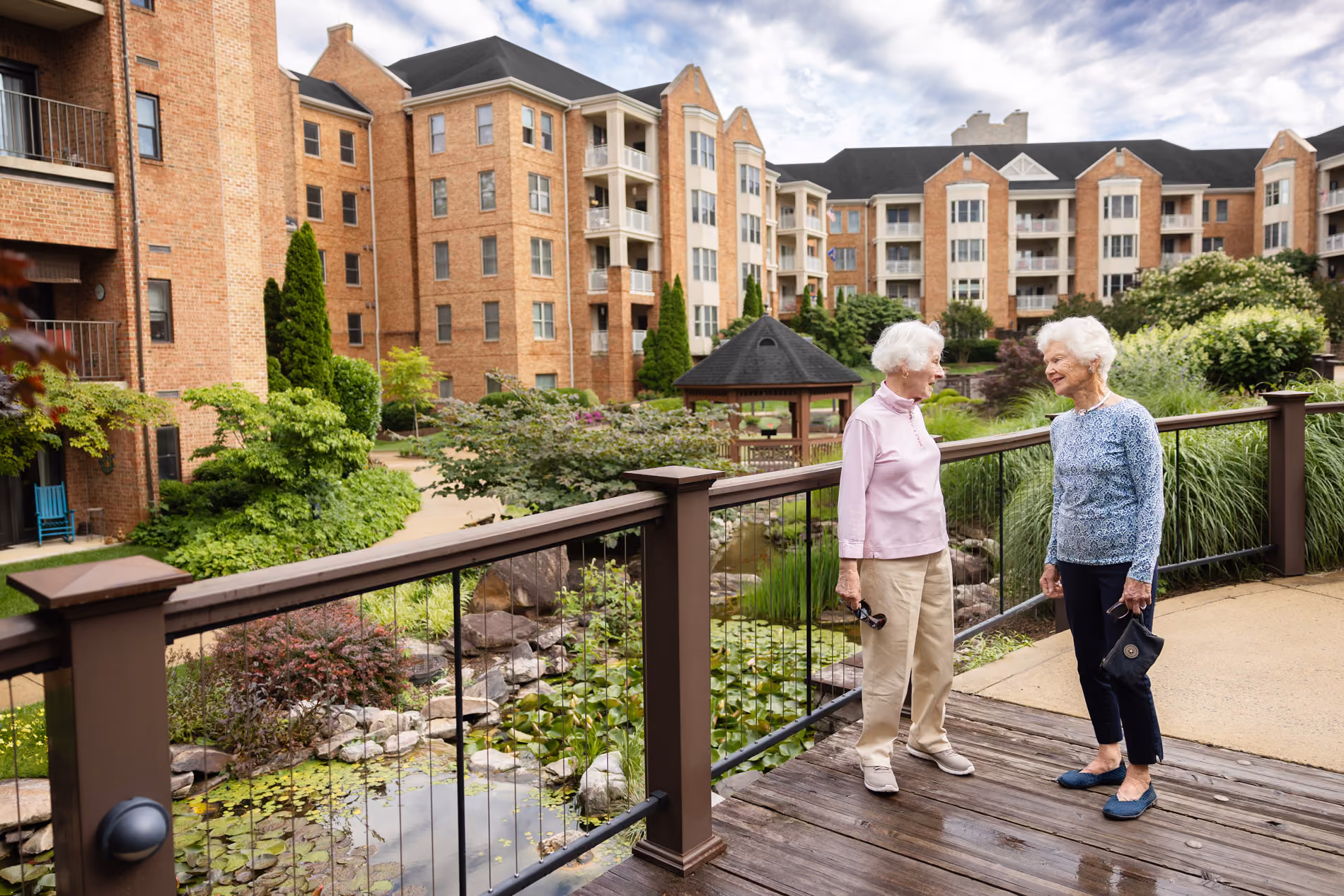 Two elderly women talking and smiling on a wooden bridge in a residential courtyard.