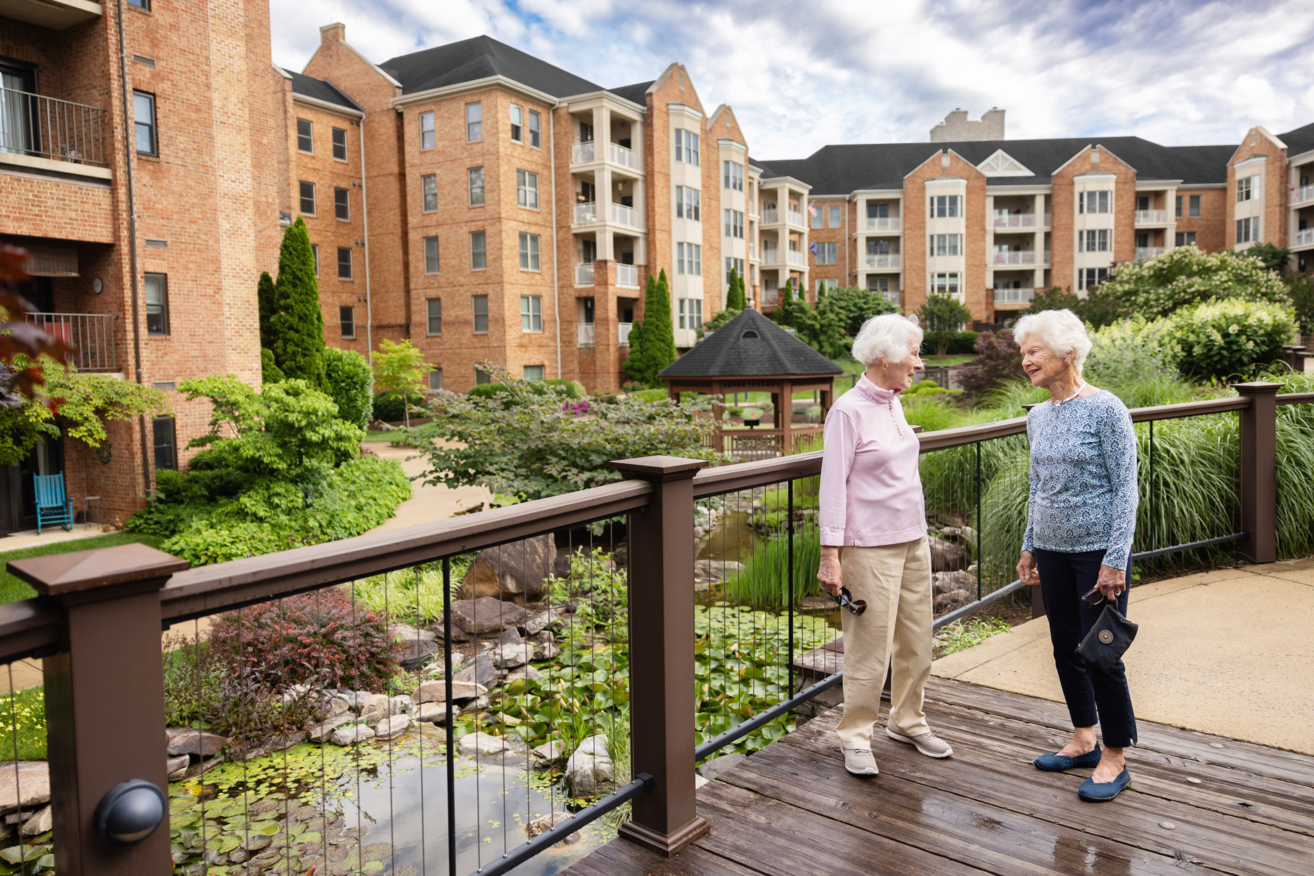 Two elderly women talking and smiling on a wooden bridge in a residential courtyard.