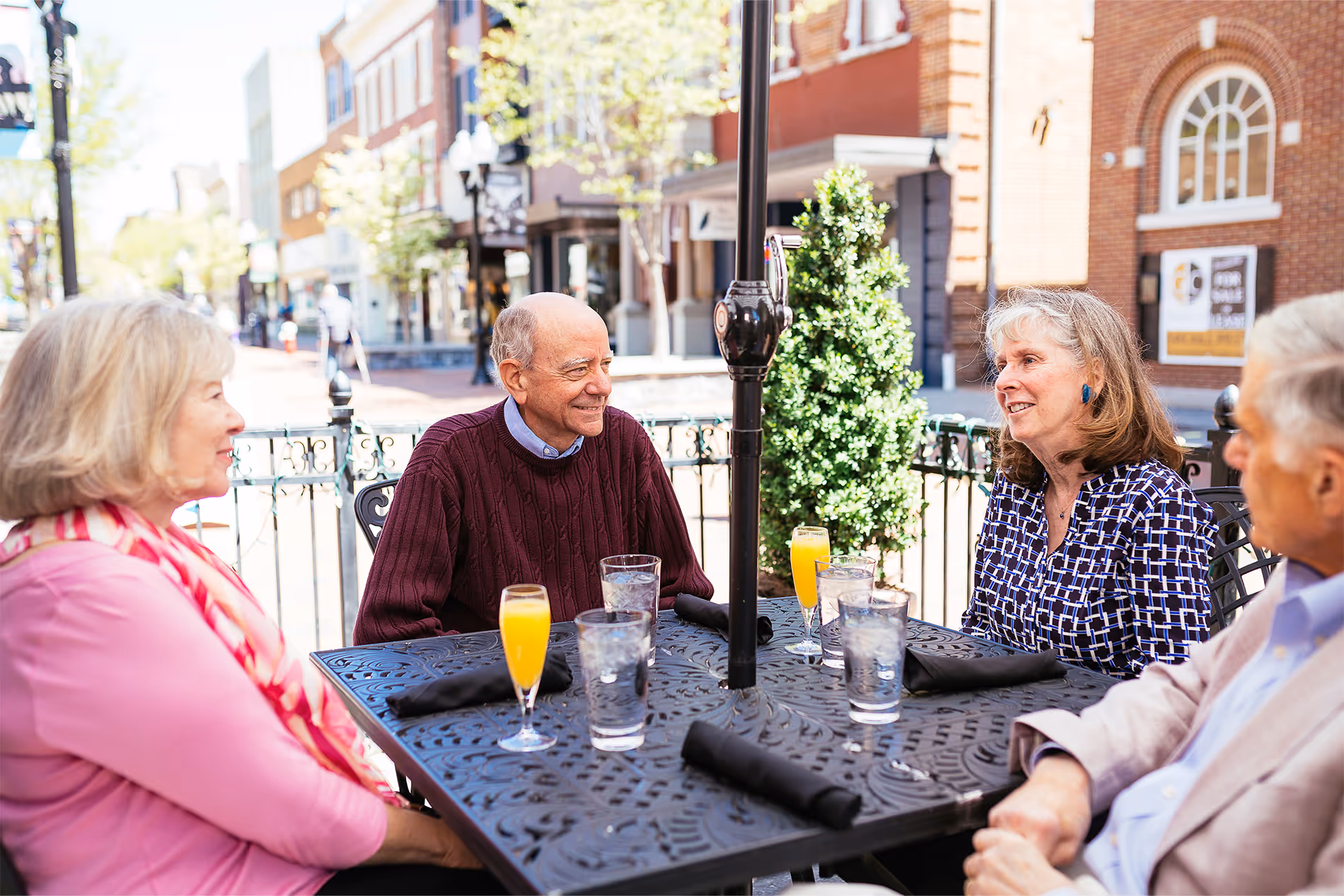 Four older adults sitting at an outdoor cafe table, enjoying drinks and having a conversation.