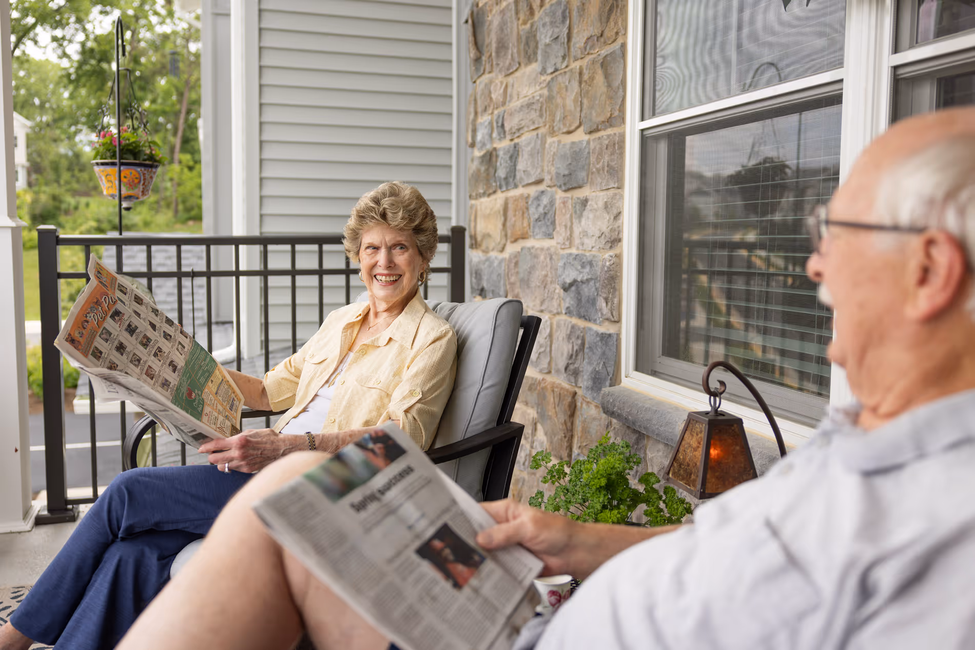 Two elderly people sitting on a porch, smiling and reading newspapers.
