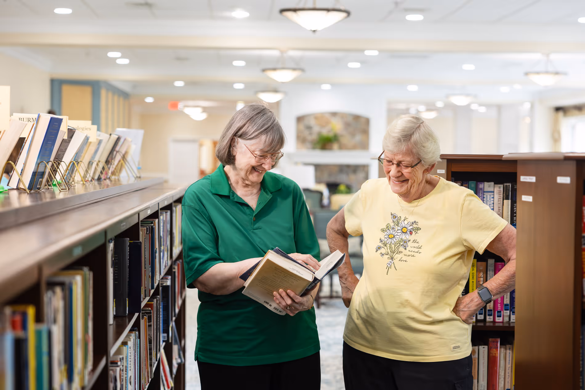 Two elderly women are standing in a library, smiling and looking at a book together.
