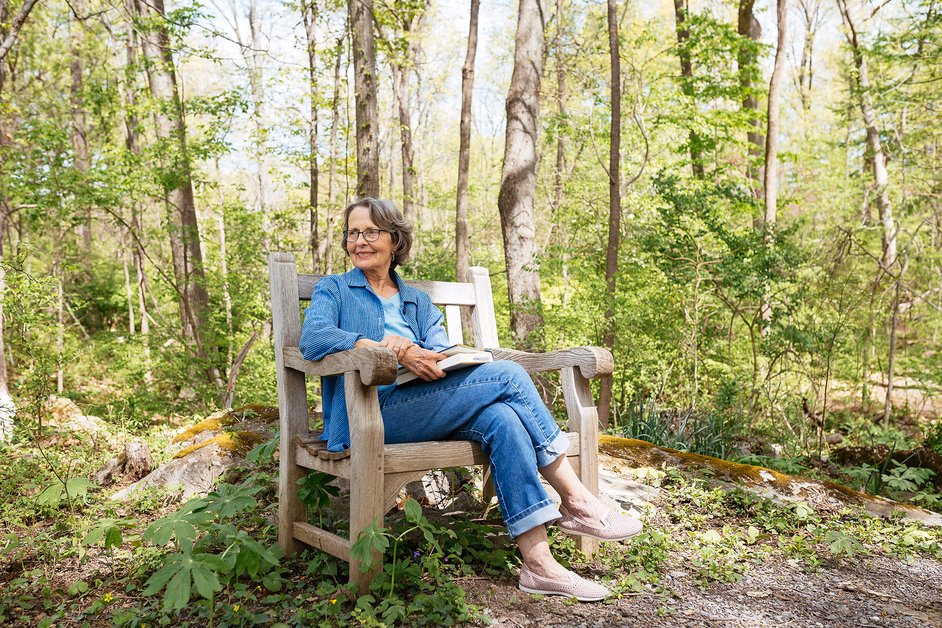 A woman sits on a wooden bench in a forest, holding a book and smiling.