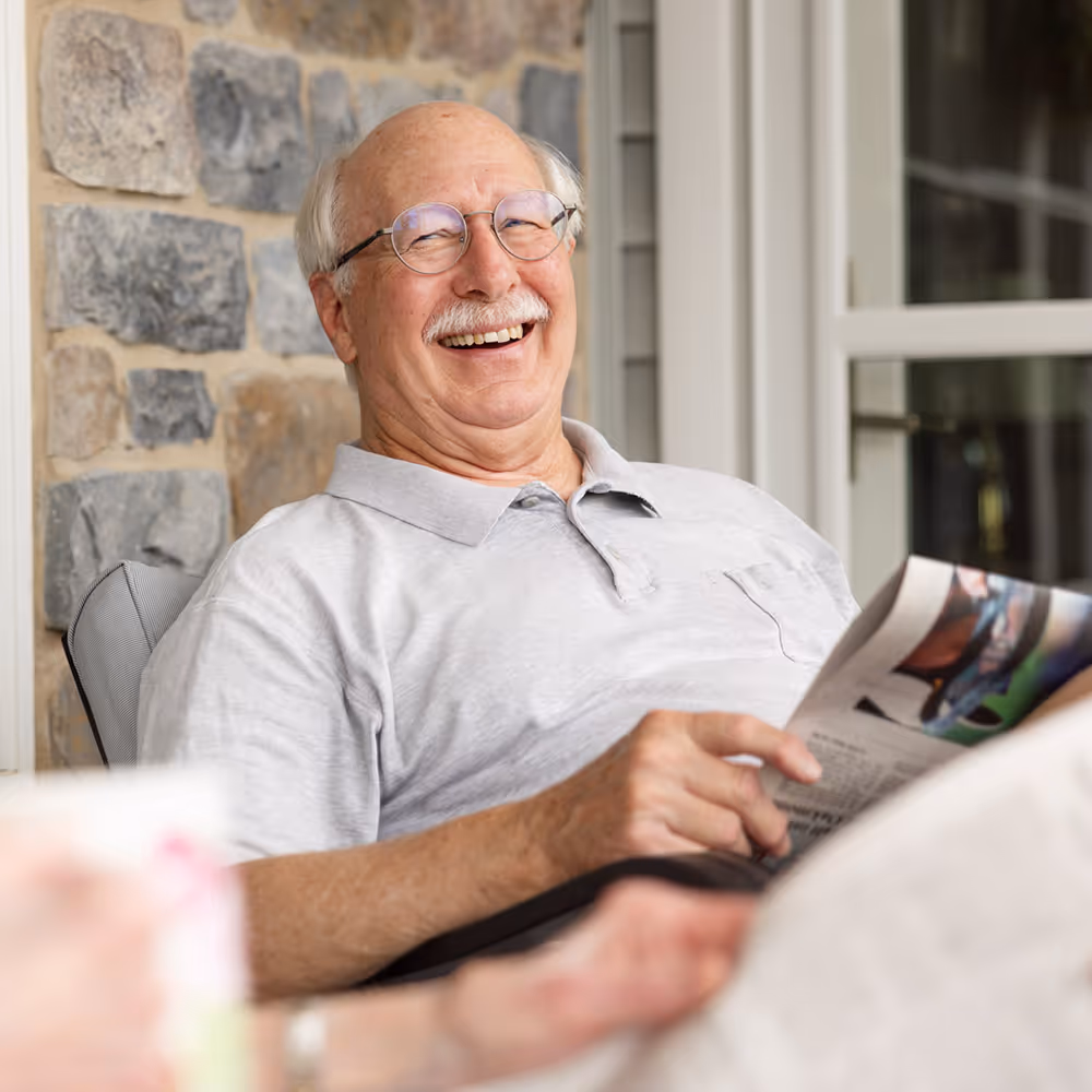 Elderly man sitting outside, smiling broadly while holding a newspaper.