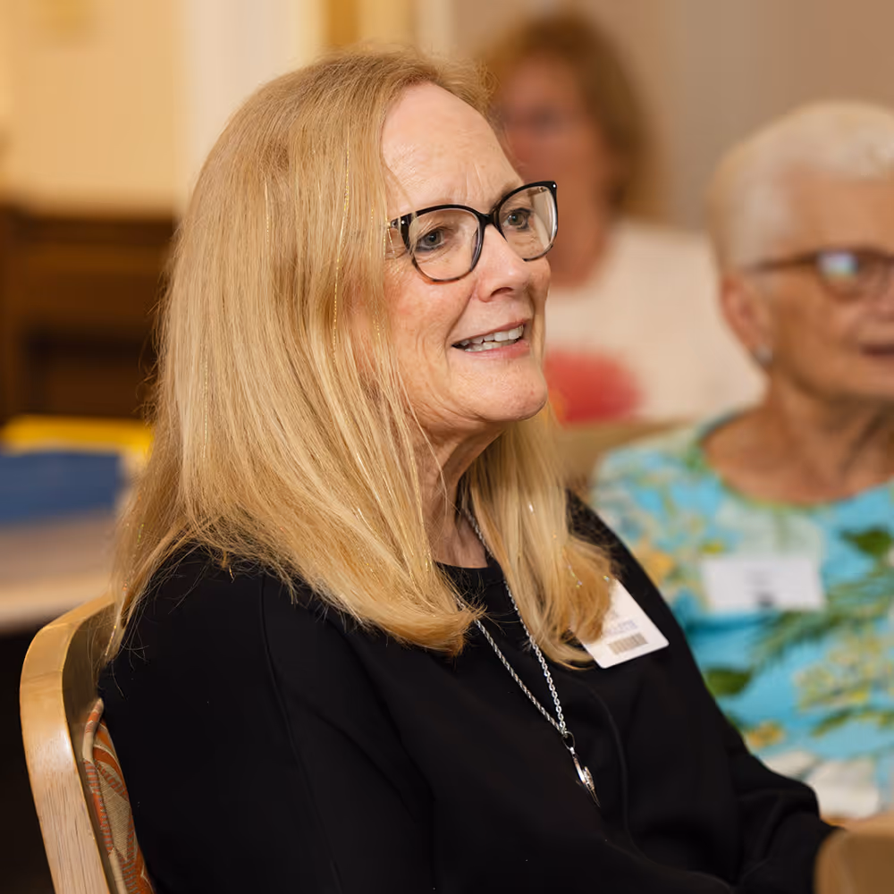 A woman with glasses and long hair is sitting and smiling during a social gathering.