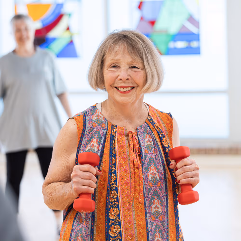 An older woman smiling while holding small red dumbbells during a fitness class indoors.