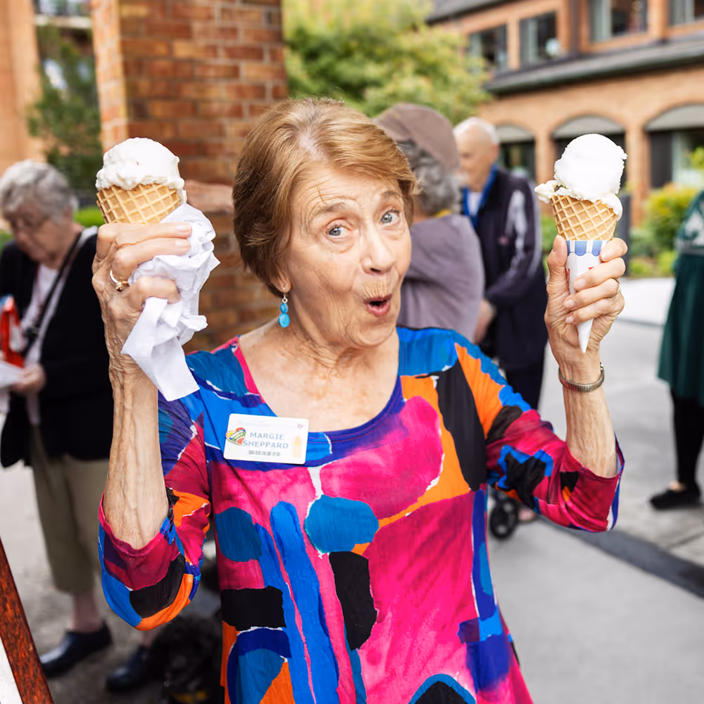 An elderly woman in a colorful top happily holds two ice cream cones outdoors.