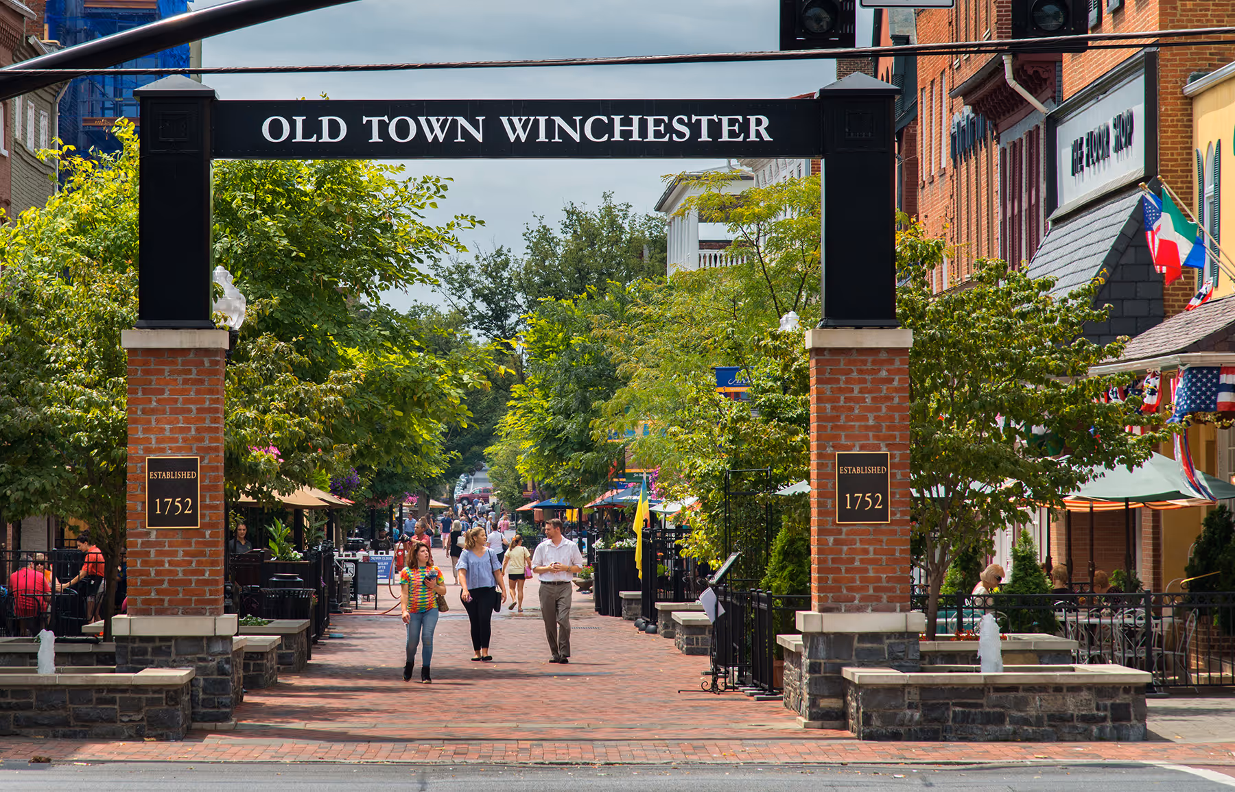 People walking on a brick path under an "Old Town Winchester" archway in a shopping area.